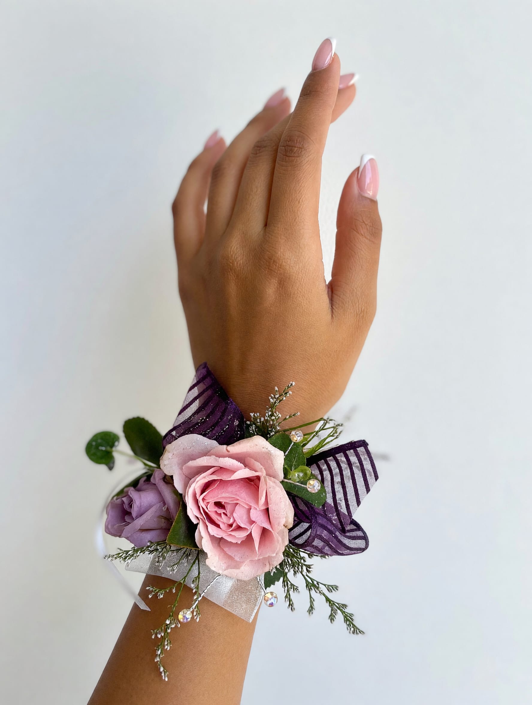 Pink &amp; Lavender Spray Rose Corsage - A beautiful wrist corsage featuring soft pink and lavender spray roses, accented with fresh greenery and finished with an elegant purple ribbon and sparkling rhinestone bracelet. Handcrafted for weddings, proms, homecoming, and other special occasions, this timeless design adds a graceful touch to any celebration.