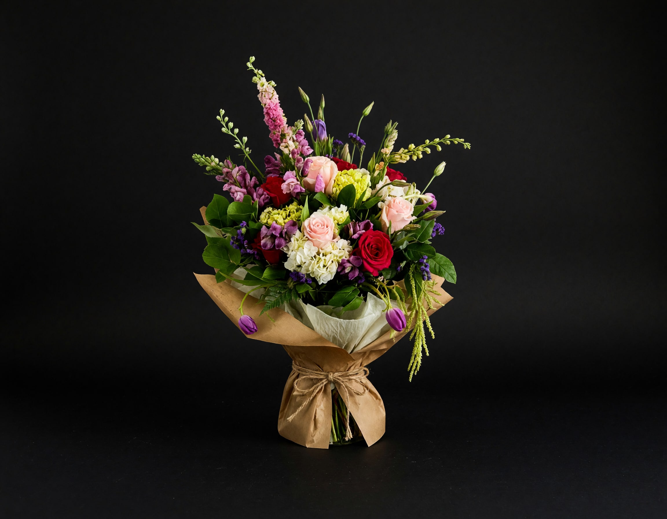 Mixed bouquet of roses, hydrangeas, and purple blooms wrapped in brown paper
