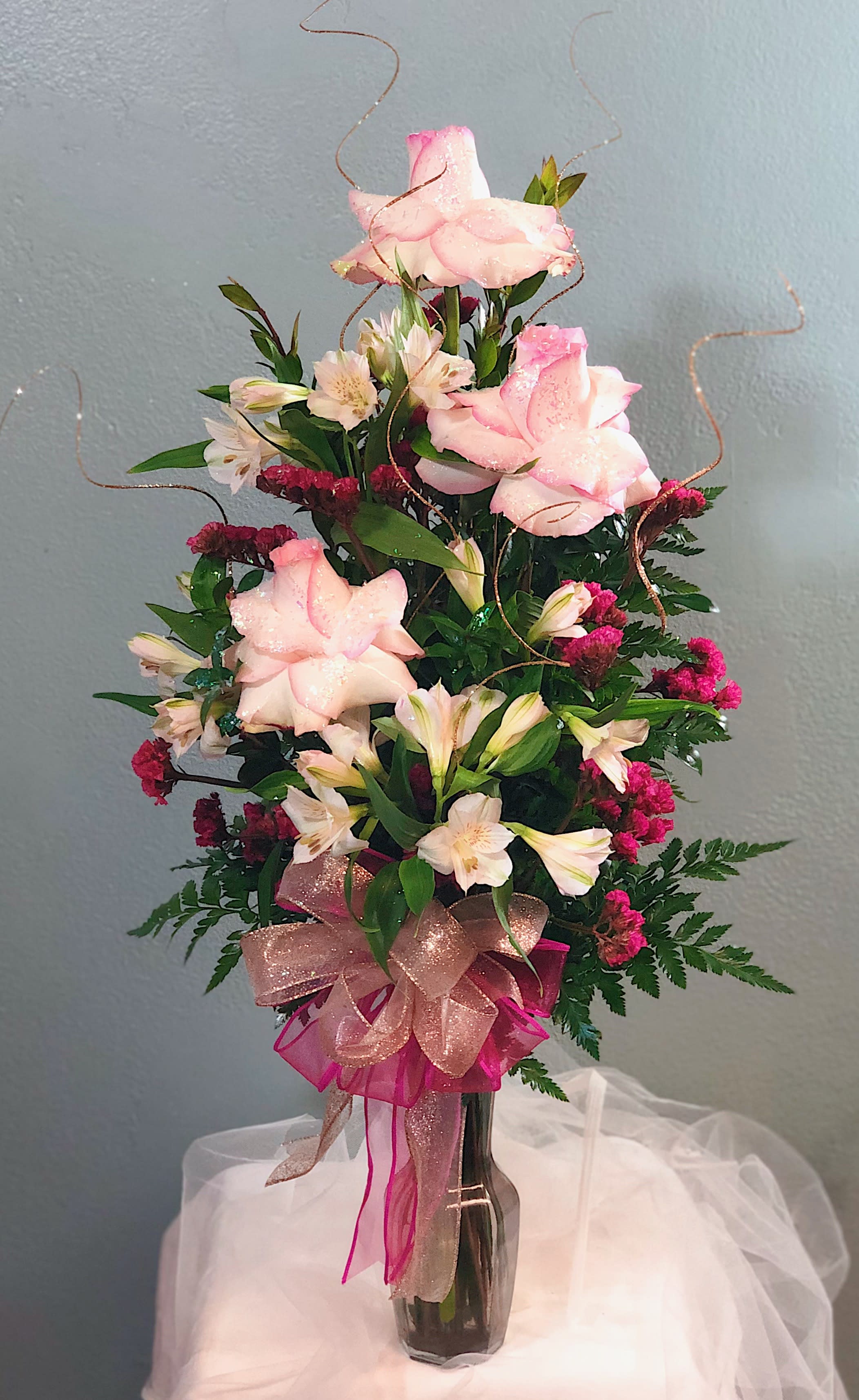 Tall pink and white rose and alstroemeria arrangement in a dark glass vase with glittery ribbons