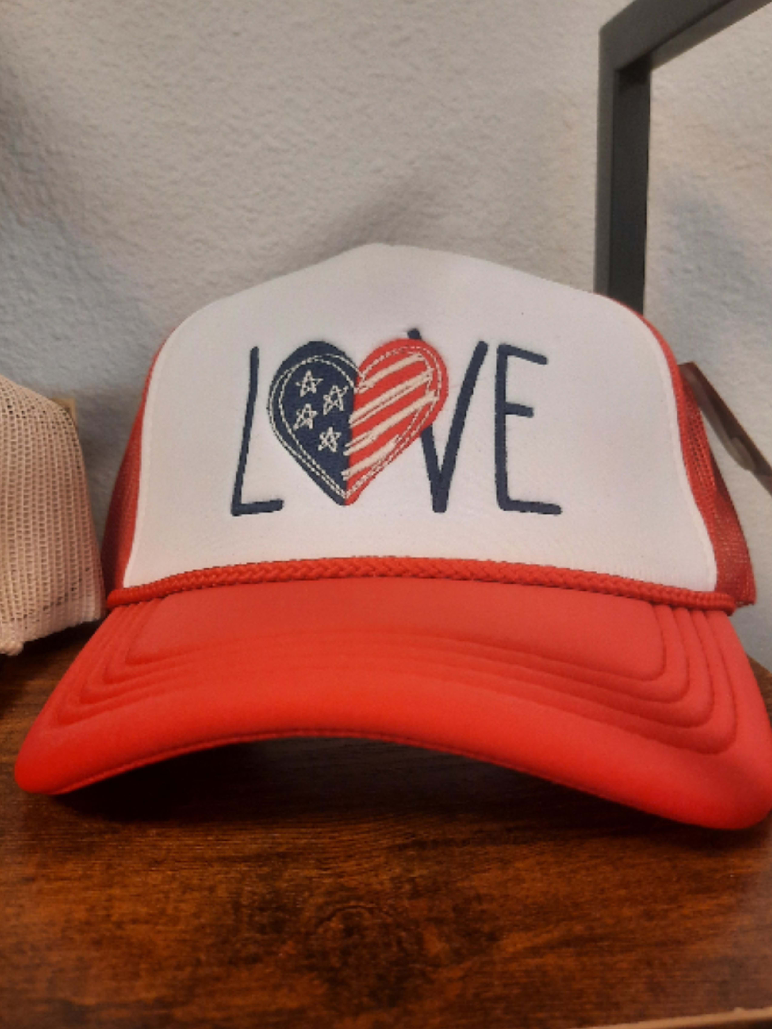 Love Trucker Hat-RED - A playful red-and-white trucker hat that reads LOVE in bold navy letters, with the O replaced by a heart in a red, white, and blue pattern - a casual, giftable accessory that works for Valentine's Day, Fourth of July, or any moment you want to send a cheerful, patriotic message. As a non-floral gift option from our collection, it gives the person you're thinking of a wearable keepsake that feels both festive and relaxed.  The hat features a curved red brim, a solid front panel, and a breathable white mesh back for comfortable all-day wear. The graphic heart detail combines classic Valentine warmth with stars-and-stripes energy, making it an easy match for red, white, and blue outfits or layered with a bouquet for a themed gift. Suggested for someone who prefers playful, practical presents-include it with a mixed bouquet for a coordinated look or order alone as a thoughtful, wearable token.