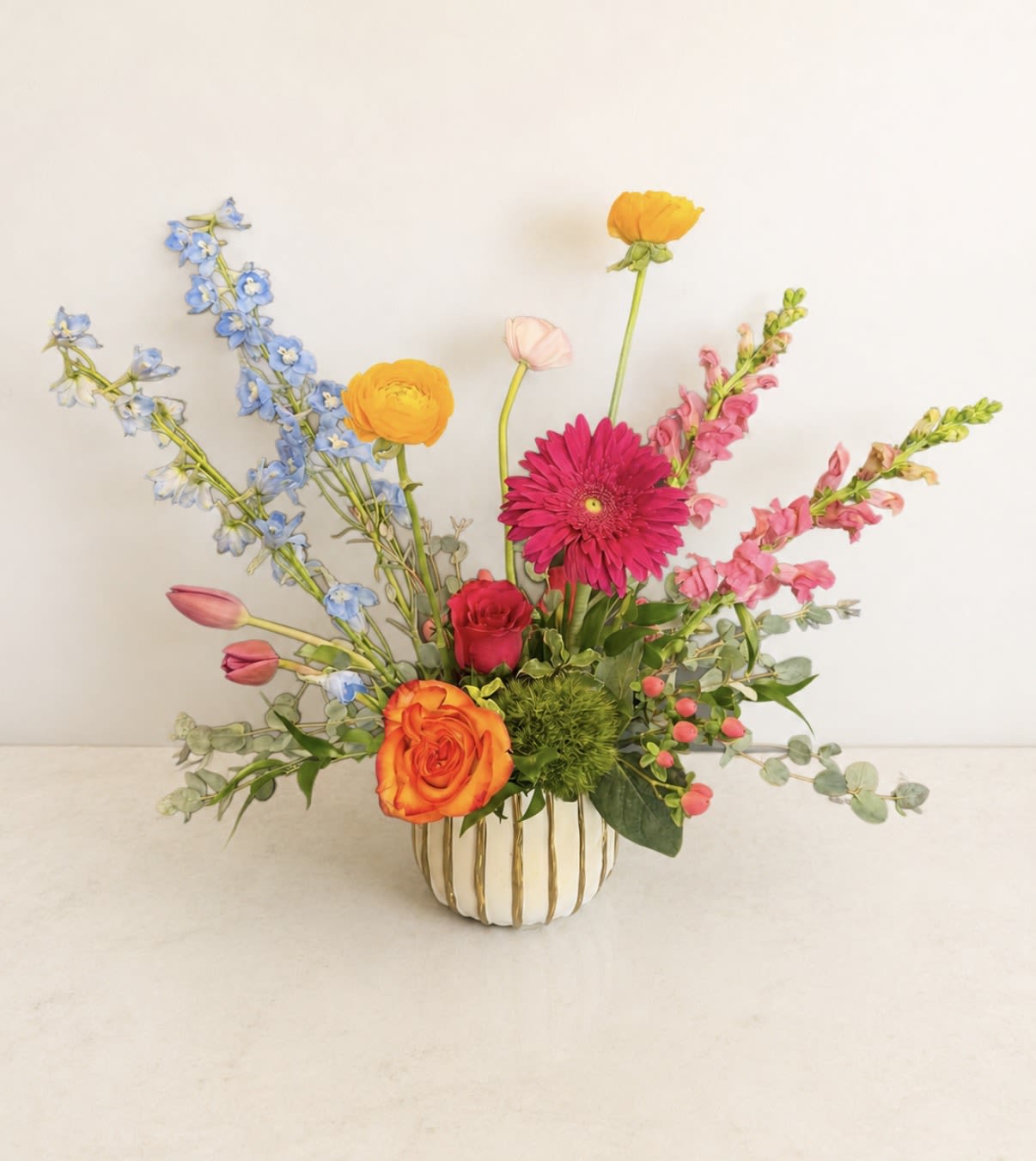 Garden Rhapsody - A lively burst of color, Garden Rhapsody is a cheerful flower arrangement perfect for a birthday bouquet, congratulations gift, or a "just because" surprise. The bright pinks and magenta focal blooms give an immediate joyful impact while the pops of orange and cool blue create a playful, modern contrast that will make the person you're thinking of smile.  This low garden-style arrangement combines roses, tulips, snapdragons, gerbera daisies, and airy blue delphinium-like stems in a loose, fanned composition. Soft blush accents and fresh green foliage add texture and balance. The flowers sit in a low ceramic vase with subtle vertical gold striping, making it ready to display on a table, desk, or mantel.  Send Garden Rhapsody to mark everyday milestones or to lift someone's spirits-the vibrant palette reads like celebration. Designed for immediate enjoyment, the arrangement arrives arranged and ready to display; fresh water and basic cut-flower care will help blooms last longer.