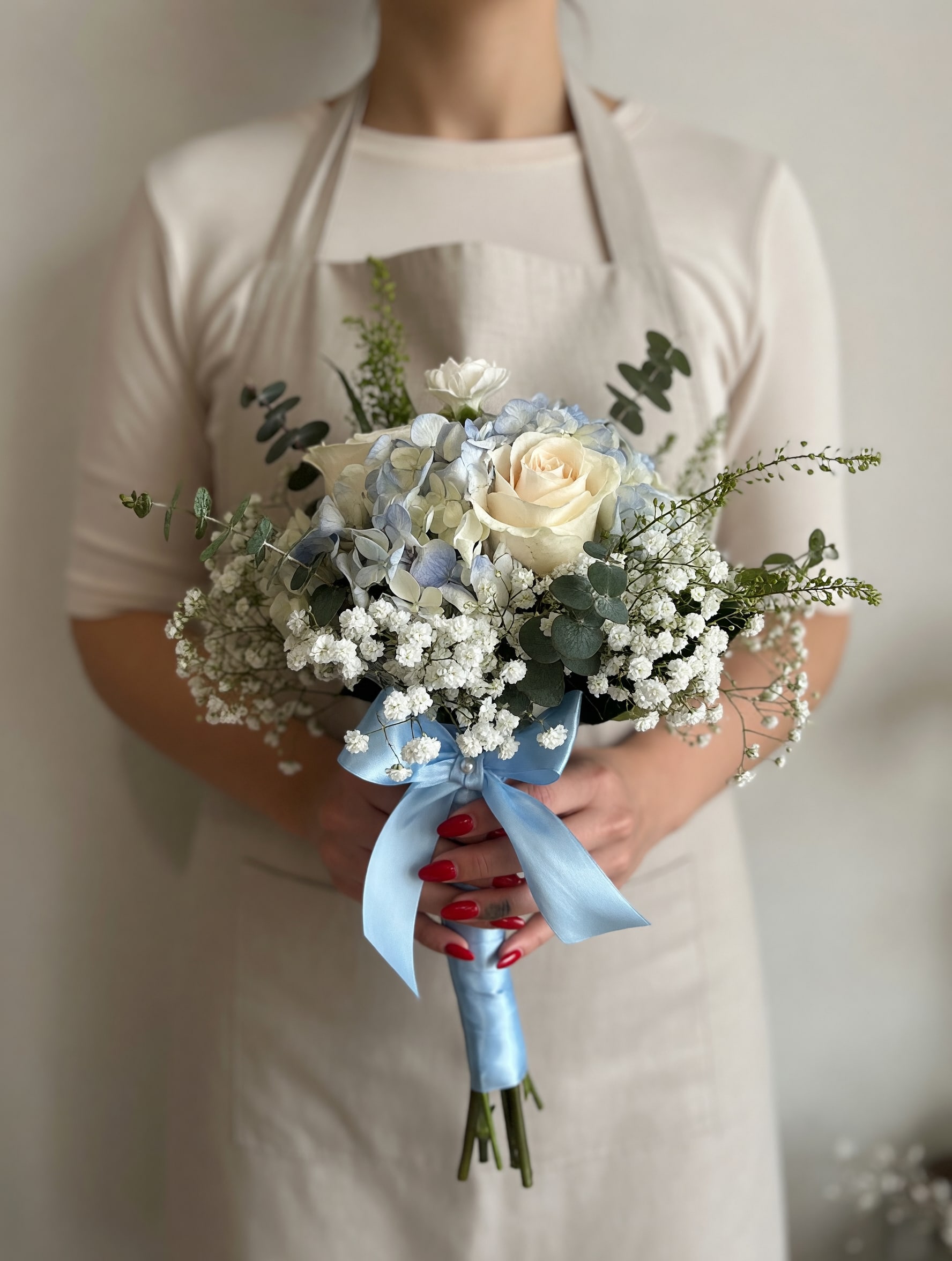 Mini Prom Bouquet Blue &amp; White  - Creamy white roses paired with pale blue hydrangeas create a soft, romantic mini prom bouquet that reads as timeless and gentle-perfect for prom, weddings, anniversaries, or Mother's Day. Airy clusters of baby's breath and silvery eucalyptus give the arrangement a light, garden-style silhouette so the person you're celebrating will feel both cherished and effortlessly graceful.  This compact, rounded bouquet mixes white, blue, and green tones with a polished finish: roses, hydrangeas, baby's breath, eucalyptus and fine filler stems are bound by a pale blue satin ribbon and presented with a neat apron wrap. The size is intentionally petite for comfortable carrying while still feeling full and lush; the satin ribbon adds a refined touch that dresses up formalwear or bridesmaid attire.  Choose this bouquet when you want someone to feel beautifully seen-whether marking a milestone or offering gentle sympathy. For best longevity, keep stems in water until the moment of use and mist lightly to preserve the petals' freshness.