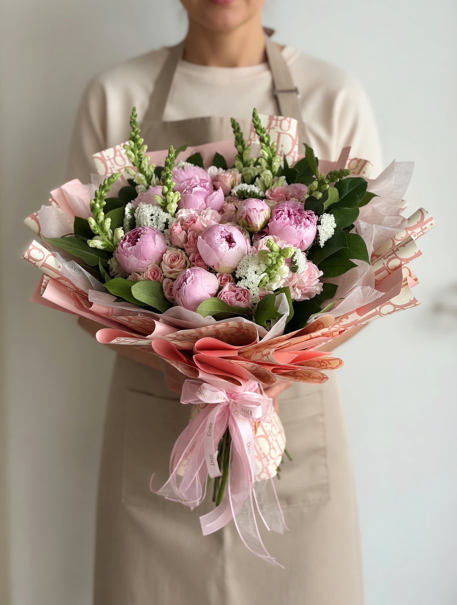 Peonies &amp; Snapdragon Bouquet - A lush mix of blush peonies, soft pink roses, and upright snapdragon spikes creates a romantic flower arrangement perfect for a birthday bouquet, anniversary roses alternative, or a thoughtful thank-you gift. The rounded peonies and smaller rose buds give a plush, tactile look while the snapdragons add vertical elegance-she'll feel cherished and seen the moment the wrapping is lifted.  Creamy white filler blooms and broad dark green foliage add depth and contrast, and the palette of pink, peach, white, and green keeps the design fresh and feminine. Wrapped in patterned pink-toned paper and finished with a sheer pink ribbon, the presentation is polished and gift-ready without extra fuss. Ideal for everyday gifting or special occasions, stems arrive arranged and ready to display; keep flowers in fresh water, away from direct heat, and trim stems for best longevity.