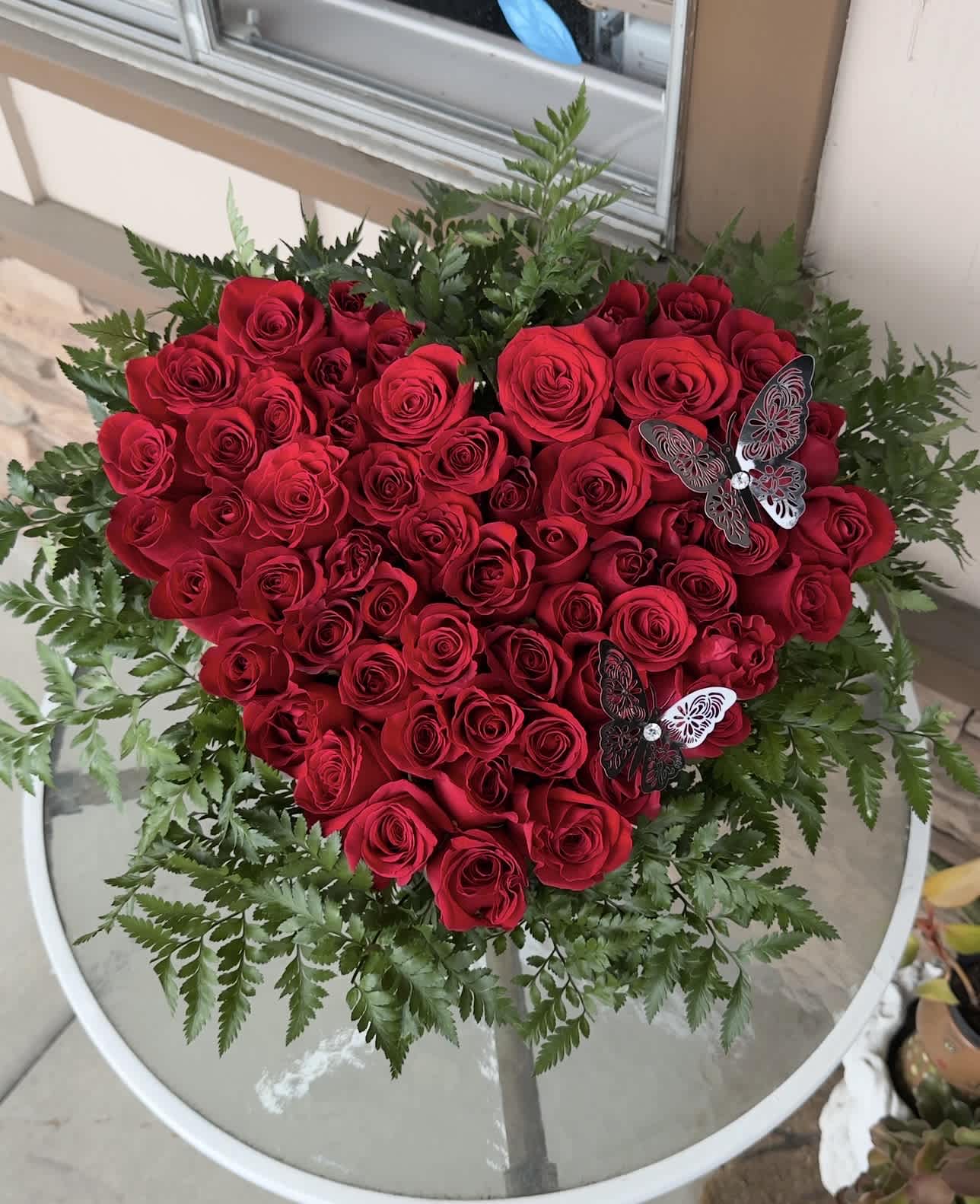 Heart-shaped bouquet of red roses with fern-like greenery and butterfly accents