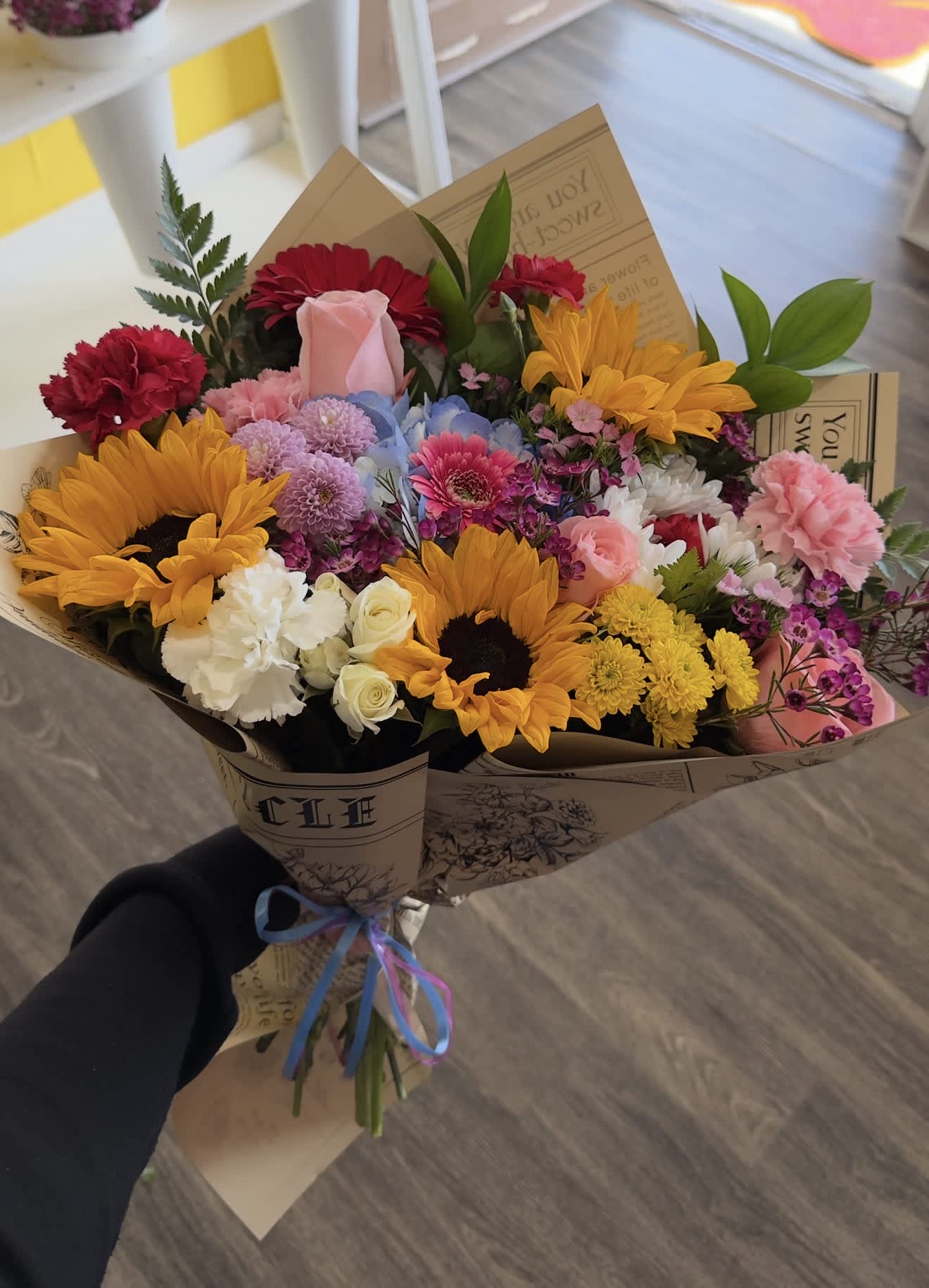 Handheld bouquet with sunflowers, roses, carnations, and mixed blooms in paper wrap