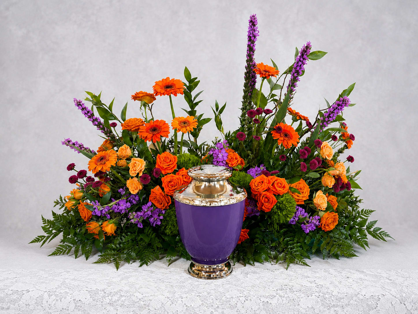 Vibrant Memories Urn Tribute - Bright, ceremonial and full of presence, this sympathy urn tribute centers on a purple urn vase for a bold memorial display. The Vibrant Memories Urn Tribute is crafted as a large, low arrangement meant for funerals or celebration-of-life services, and its orange, purple, pink and green palette creates a vivid complementary contrast that draws the eye and honors a life with color.  The design mixes bright orange roses and gerbera daisies with purple accent flowers, chrysanthemums and small burgundy blooms arranged broadly around the central urn. Tall spires rise from the back to add height and formality while the spread of blooms keeps the overall silhouette low and substantial. Textural greens fill and soften the composition, giving depth and a natural finish.  Perfect for family memorials or a service where a striking, colorful tribute is desired, this piece communicates warmth, energy and thoughtful remembrance so the person you're honoring feels truly seen. Size and scale make it suitable for placement on a casket spray alternative table or memorial display; standard care is to keep flowers in a cool location and away from direct heat for best longevity.