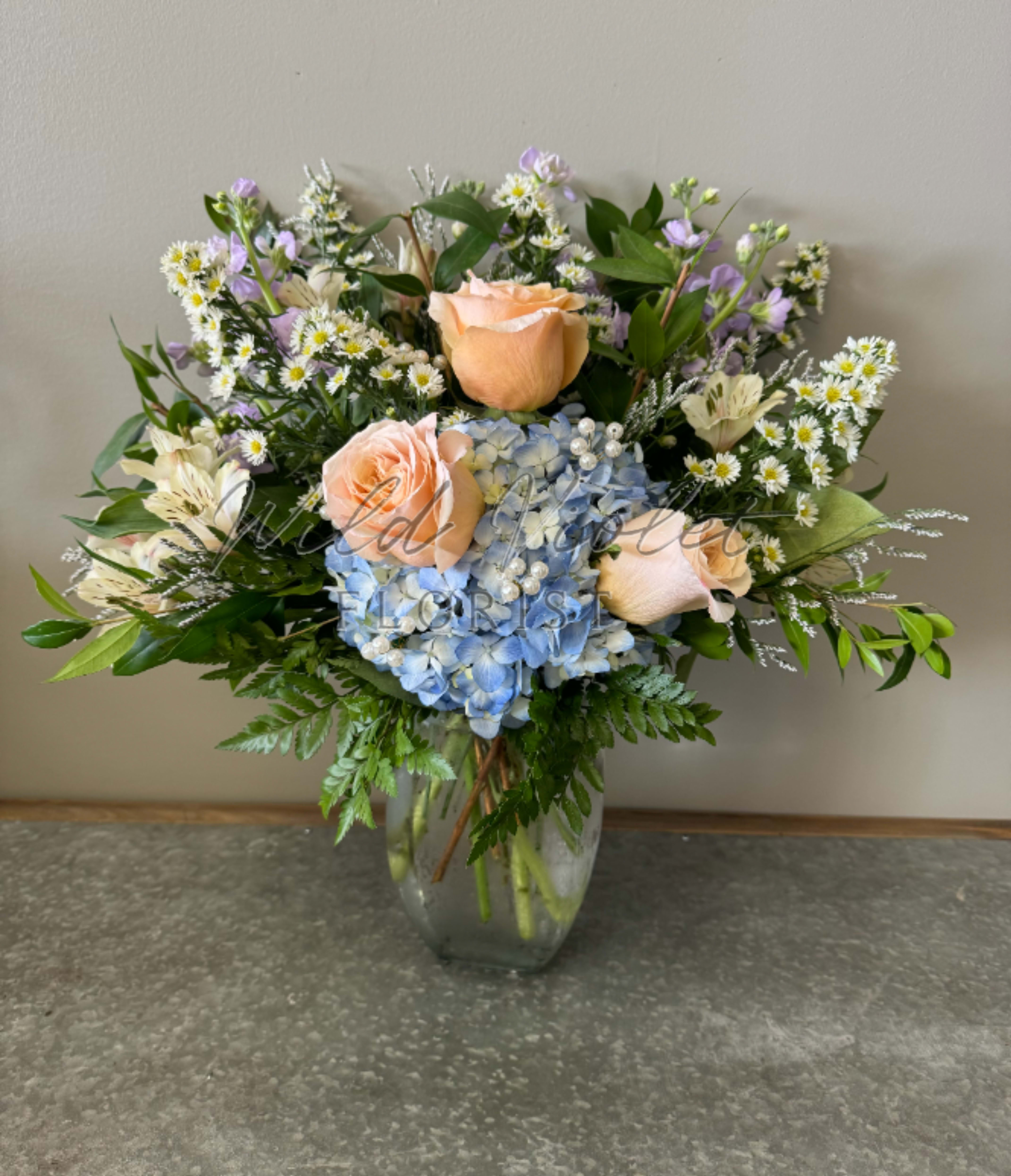Mixed bouquet of peach roses, blue hydrangea, and small white daisies in a glass vase