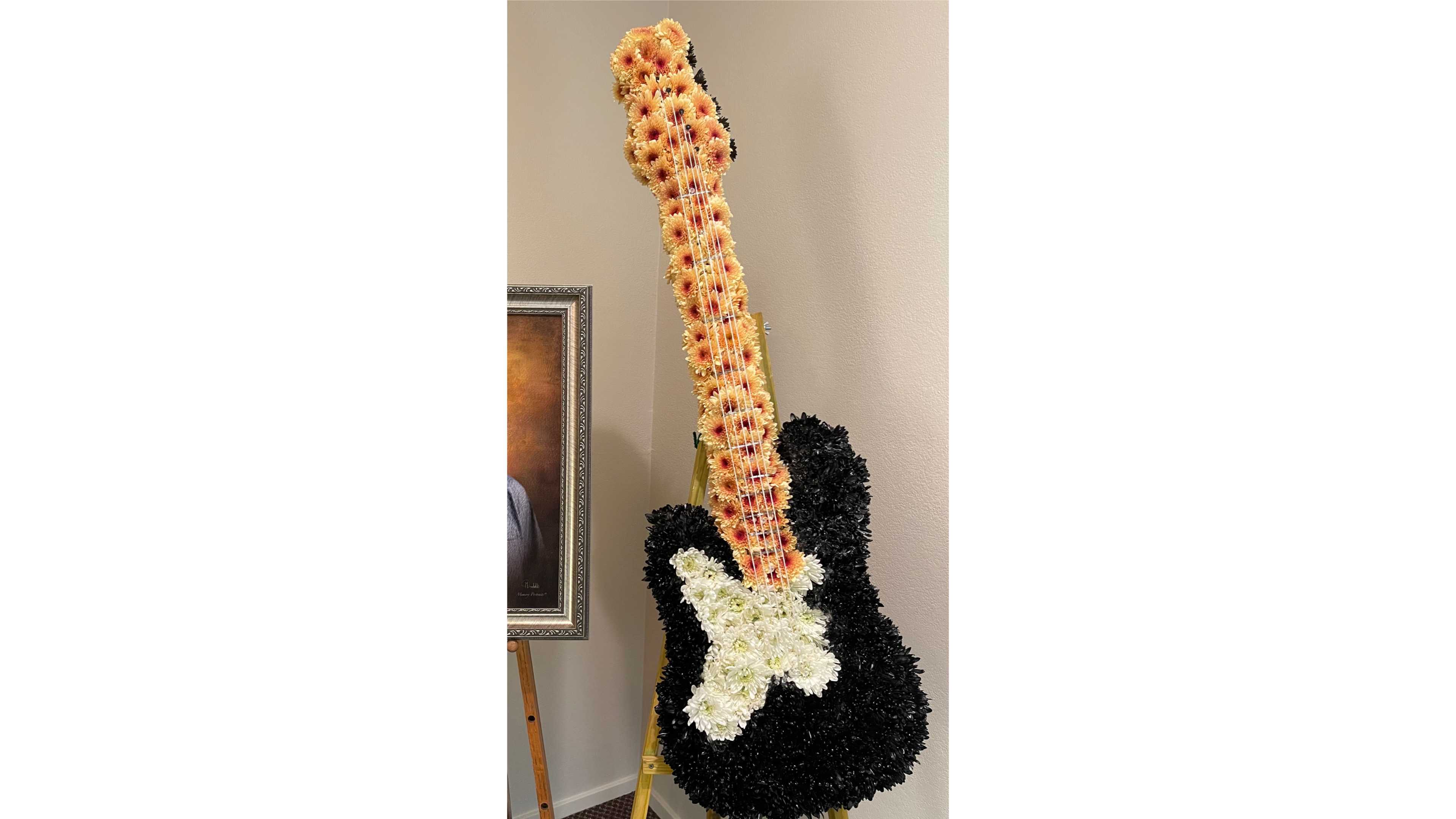Standing floral arrangement shaped like a guitar with black, peach, and white flowers on an easel.