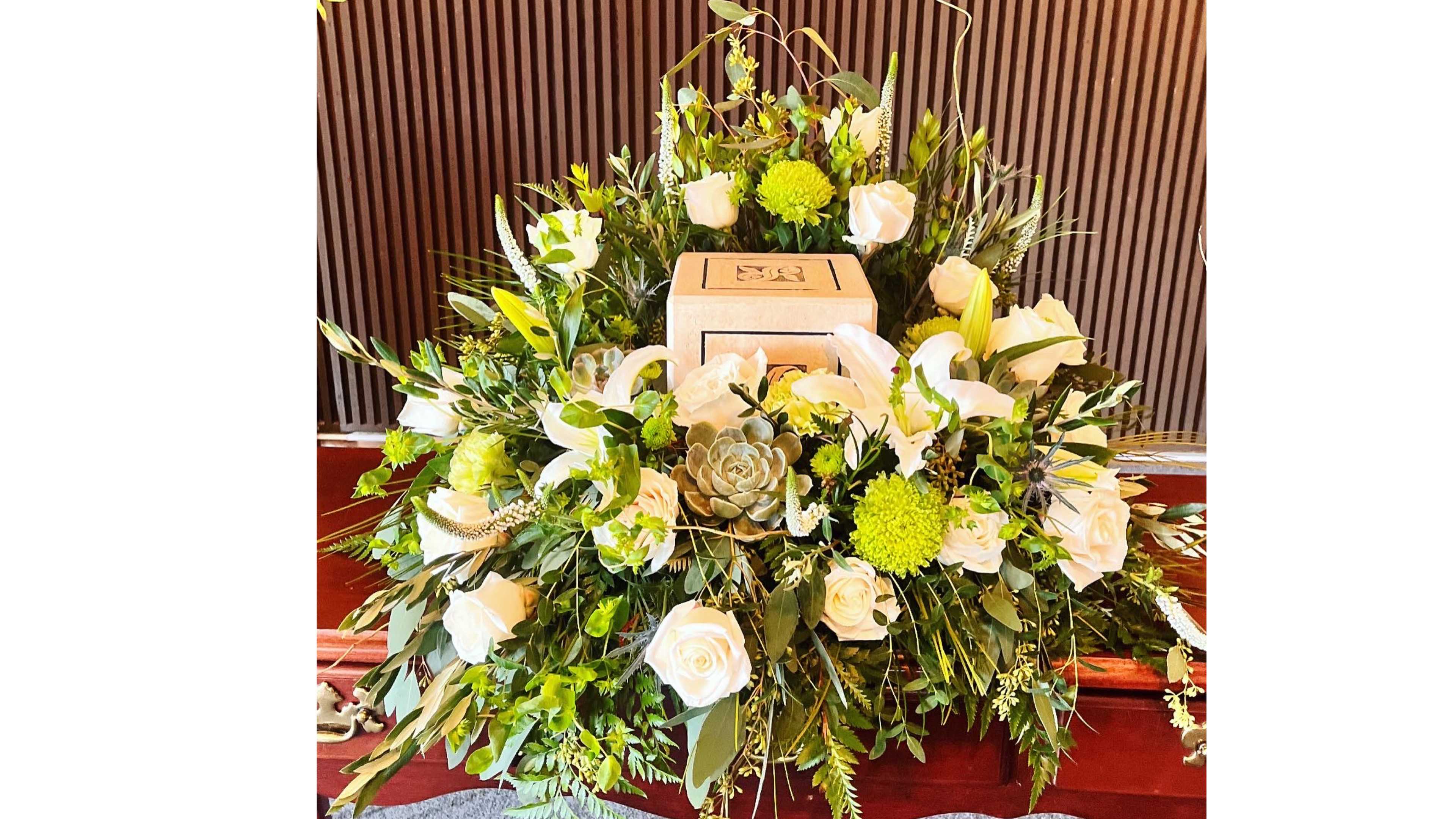 Cremation urn surrounded by white roses, lilies, green mums, and a succulent on a wooden casket