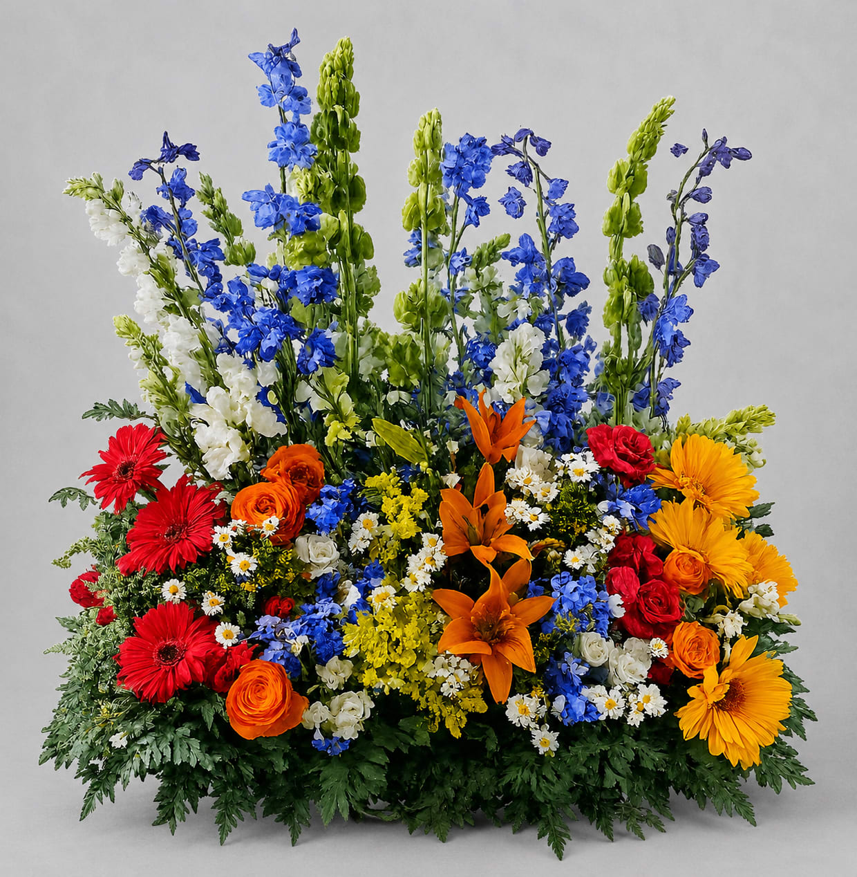 Colorful Tribute Table Arrangement  - Tall blue delphinium spires crown this Colorful Tribute Table Arrangement, giving it a striking vertical silhouette that catches the eye - perfect as a birthday bouquet, celebratory sympathy table piece, or a cheerful congratulations gift. The bold mix of mixed color blooms reads joyful and garden-fresh, so the person you're honoring will feel uplifted and truly seen by this lively floral statement.  The lower, broad base is densely packed with bright red gerbera daisies, vibrant orange roses and ranunculus, sunny yellow daisy-like blooms, and clusters of small white daisies for contrast, creating a layered, textured look with lots of movement. The combination of lilies and other seasonal flowers adds depth and scent, while the tall green accents bridge the composition for a full, abundant presentation. Sized to make an impact on a tribute table or as a centerpiece, it brings a festive, everyday elegance without feeling fussy.  Ideal for celebrating life, offering warm thanks, or bringing color to a gathering, this arrangement reads as both heartfelt and uplifting for the recipient. Order early for special dates to ensure availability; the flowers arrive arranged and ready to display, with simple care to keep blooms fresh - fresh water, a cool spot away from direct sun, and daily water checks will help the recipient enjoy it longer.