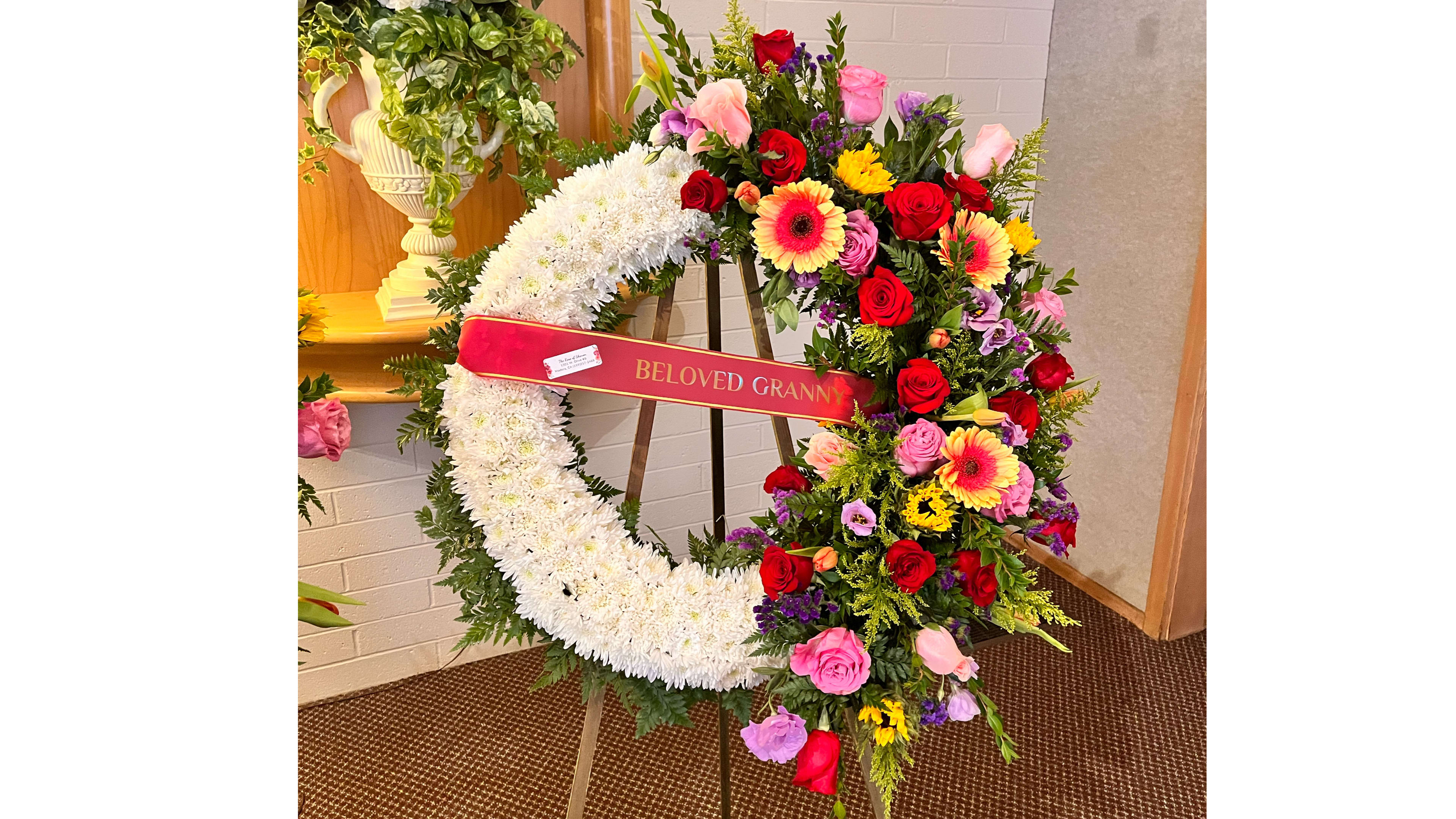 Standing white sympathy wreath with bright mixed flowers and a 'Beloved Granny' ribbon on an easel