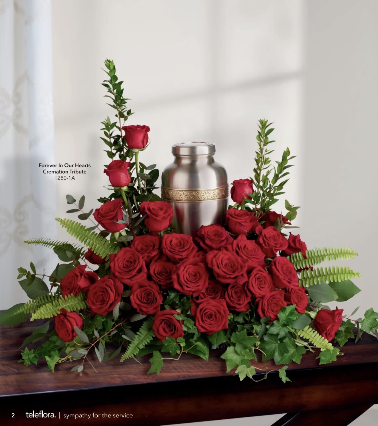 Forever In Our Hearts Cremation - A dense halo of deep red roses encircles a polished silver cremation urn in this sympathy arrangement, creating a focused, reverent tribute for a funeral service. The design-low and expansive at the base with upward-sweeping accents on either side-frames the urn so the person you're remembering feels honored and held. This piece is suited for memorials and cremation services where a formal, respectful presence is needed.  Rich red roses and spray roses are paired with upright textural accents and trailing greenery in tones of green and white to soften the composition and add visual depth. The metal cremation urn sits centered on a wooden table in the display, the polished finish contrasting with velvety petals for a dignified, tactile effect. The arrangement's balanced silhouette and restrained color palette read as a classic, solemn tribute.  Choose this piece to convey enduring love and respect-the kind of memorial that helps family members feel seen and comforted during a service. Designed for immediate presentation, it arrives ready to place on the memorial table; light misting of the flowers and keeping them in a cool, sheltered location will preserve the blooms through the day.