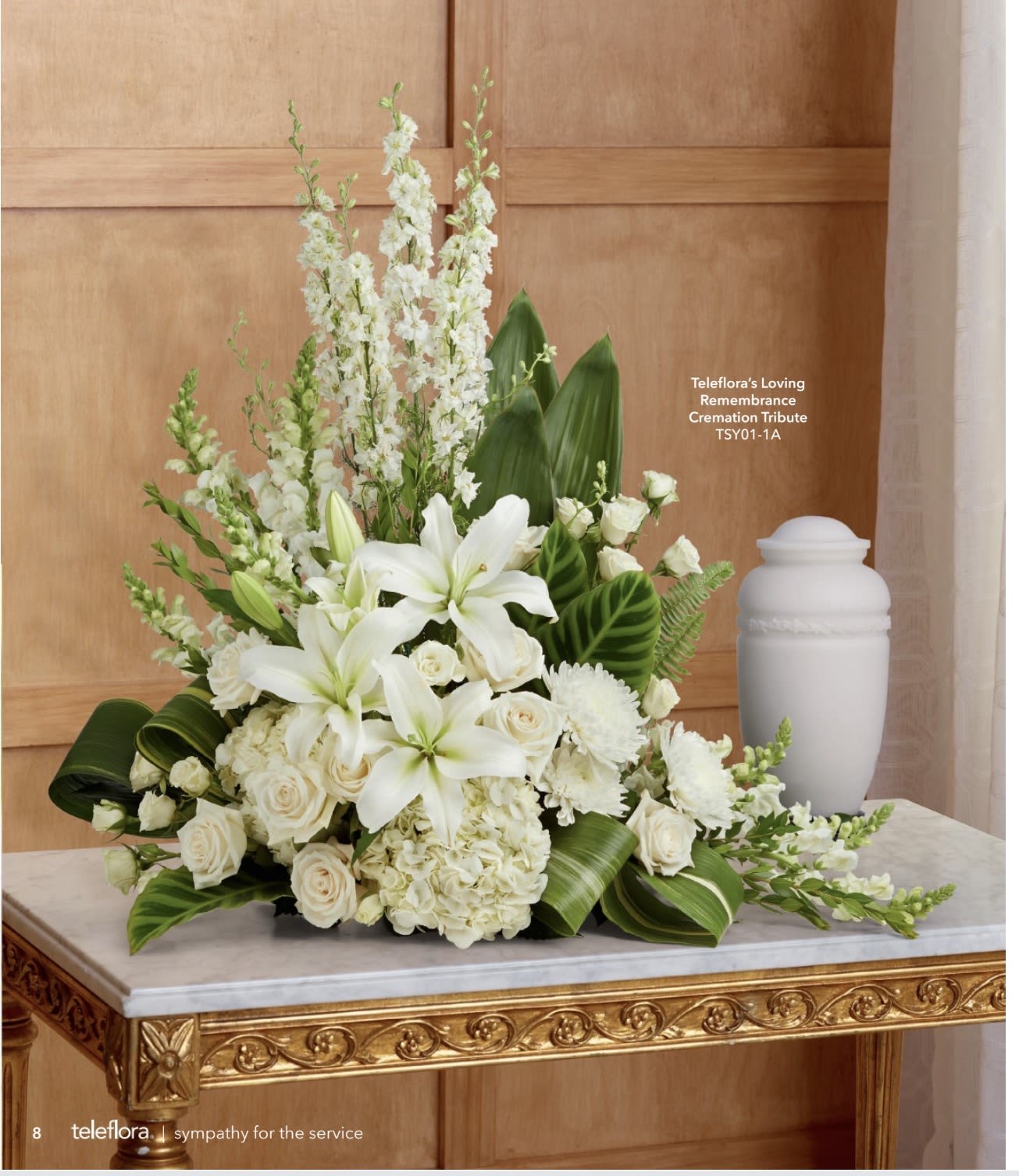 Loving Remembrance Cremation Tribute - An elegant, serene tribute built around layered white lilies and roses, this cremation sympathy arrangement brings a quiet grace to memorial services. Designed as an upright, full arrangement beside a white urn, the composition speaks to a reverent farewell - an appropriate sympathy or funeral piece that helps the person you're honoring feel remembered and respected.  Creamy hydrangeas and soft chrysanthemums add volume and a gentle texture, while tall snapdragon spikes provide vertical movement and a sense of uplift. Broad glossy greens frame the mostly white palette, giving contrast and a living backdrop to the blooms. Sized to complement a standard cremation urn and funeral setting, this arrangement delivers a traditional, comforting look that reads well year-round.