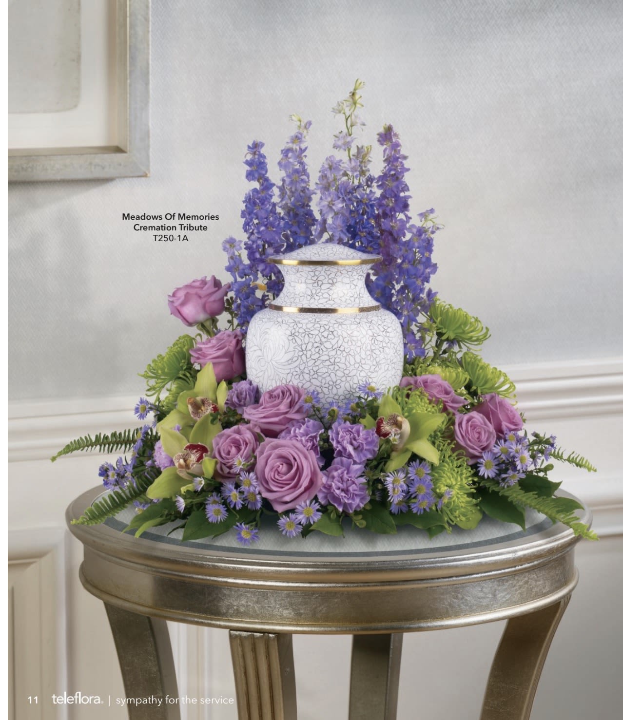 Meadows of Memories - A soft halo of lavender roses and purple carnations frames a white decorative urn in this sympathy flower arrangement, creating a serene cremation tribute designed for a service setting. Meadows of Memories is a sympathy flower arrangement that gently balances vertical movement and compact fullness so the person you're honoring feels wrapped in calm color and thoughtful design.  The design features a low ring of lavender roses and small purple daisy-like blooms paired with chartreuse-green cymbidium orchid accents and touches of pink and white blooms to soften the palette. Tall blue‑purple spike flowers rise behind the urn for layered height while the base remains lush and close to the display table, giving the piece presence without overwhelming the space. The mix of roses, orchids, daisies and carnations offers varied textures-from silky petals to crisp orchid leaves-that read peaceful and refined from every angle.  Made to sit beside an urn on a display table at a memorial or funeral service, this arrangement conveys sympathy and quiet remembrance so the person you're thinking of will feel honored and comforted. Delivered complete with the white urn and arranged for immediate presentation, care is simple-keep in a cool, shaded spot and mist sparingly if needed to maintain freshness through the service.