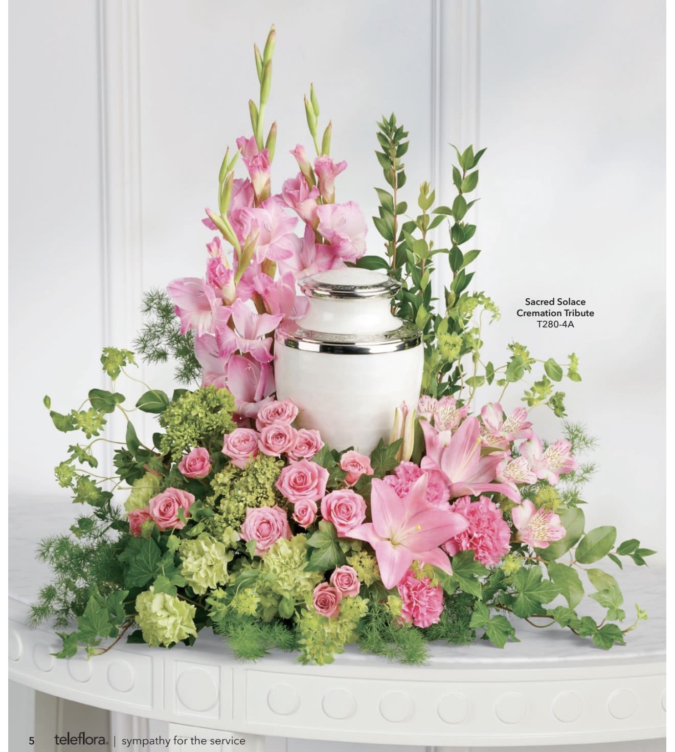Sacred Solace Cremation Tribute - A soft pastel tribute arranged around a white cremation urn, this sympathy floral spray uses pink, green, and white blooms to create a calm, reflective presence for a funeral or memorial service. The composition is designed as a cremation tribute and will help the person you're honoring feel remembered with dignity and tenderness.  Tall pink gladiolus-like spikes rise behind the urn, while pink roses, carnations, lilies, alstroemeria, hydrangea and pale green accents form a dense, layered base that gently envelops the vessel. Upright greenery and airy filler add balance and a formal, contemplative silhouette. Sized to sit gracefully with a standard urn, this arrangement arrives ready to place at a service or graveside; light misting keeps blooms fresh during the day of the ceremony.