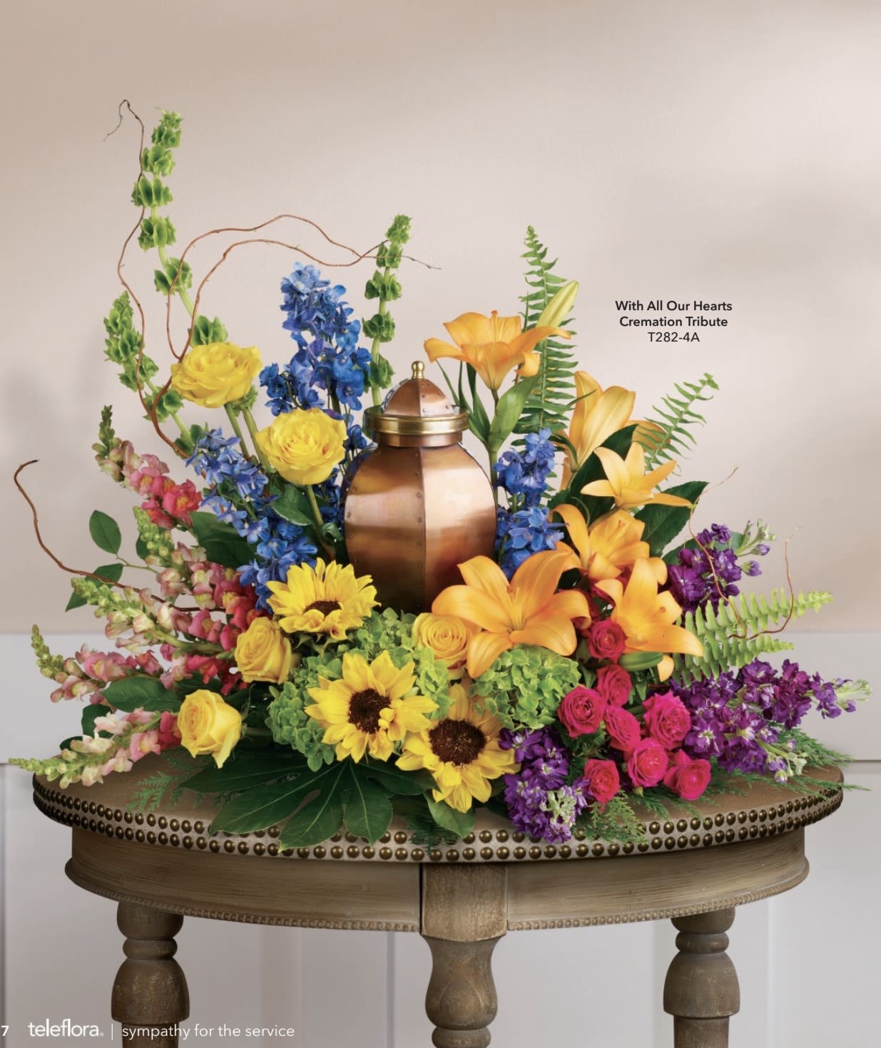 With All our Hearts Cremation - A bright, ceremonial sympathy arrangement centered on a warm copper urn, designed to honor memory with color and presence. This funeral floral piece places yellow and orange lilies and large sunflower faces around the urn, with tall blue delphinium-like spikes and pink snapdragons rising behind to give a formal, uplifting silhouette-a meaningful choice when you want the person you're remembering to stand out in the service.  The composition mixes yellow roses and spray roses with hydrangea textures and purple accents for depth, creating layered color from eye-level up to the taller vertical elements. Set low across a wooden table, the display spreads broadly so it reads well from all sides; the mix of blooms balances bold, sunny tones with cool blue and soft pink to convey warmth, comfort, and celebration of life.  Made to provide a ceremonial focal point at a memorial or funeral, this piece suits families seeking a vivid, dignified tribute that feels heartfelt and personal. Care and placement on a stable surface is recommended to showcase the urn-centered design; please contact us with timing or personalization requests so we can ensure delivery and presentation meet the needs of the service.