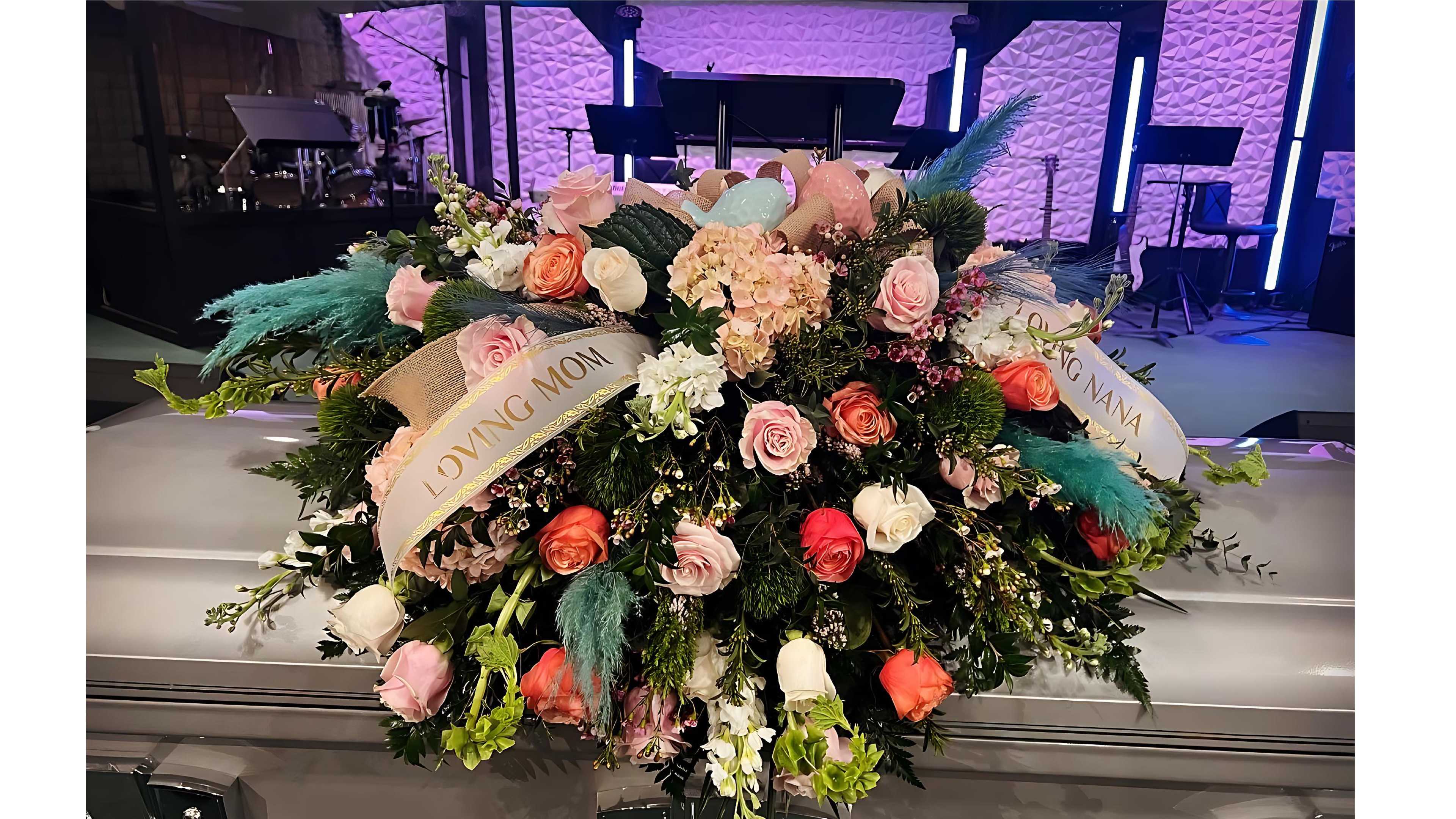 Large casket spray of pink, peach, and white roses with ribbons reading Loving Mom and Loving Nana.