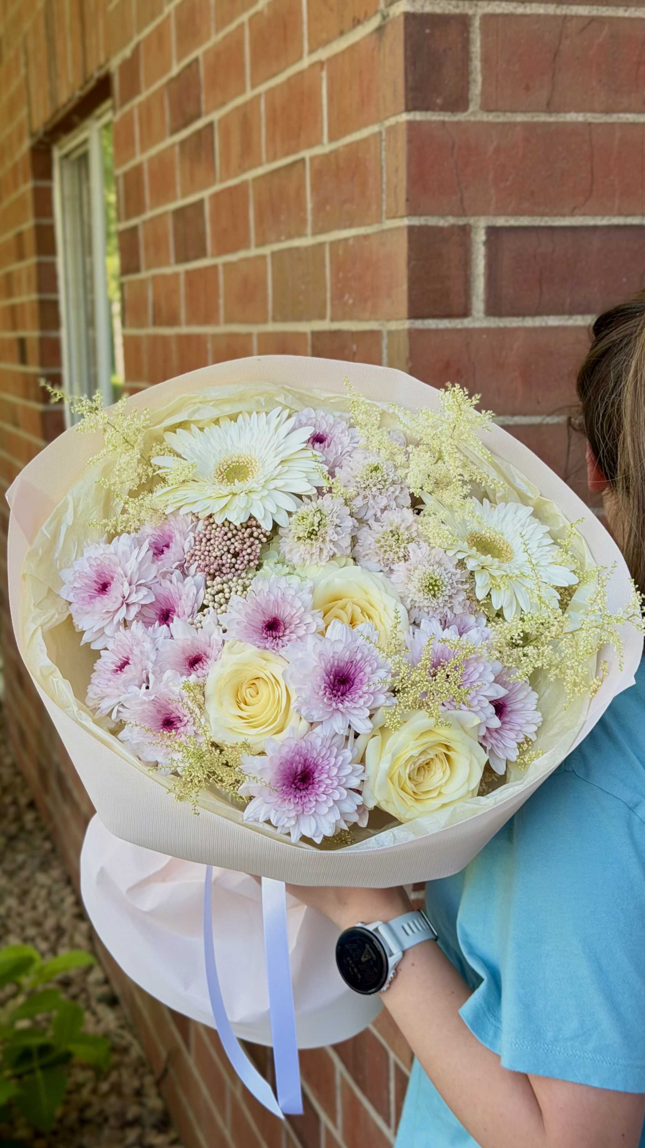 Golden Hour Garden  - Creamy pale yellow and soft pink roses mingle with lavender and blush daisy-like blooms in a hand-tied bouquet that reads like a gentle spring sunrise - a perfect birthday bouquet, anniversary gift, or bridal bouquet for someone who loves romantic pastels. The layered white and blush paper wrap with a pale ribbon gives a clean, gift-ready presentation that makes the recipient feel truly celebrated the moment they see it.  The arrangement is full and rounded, tightly clustered with cream roses, pale pink and lavender chrysanthemums and daisies, plus fine airy filler flowers that add a cloudlike texture without hiding the main blooms. Wrapped in layered paper and finished with a soft ribbon, it's designed to feel lush yet delicate, with subtle lavender centers that provide a soft contrast against ivory petals.  Ideal for weddings, anniversaries, sympathy, or a thoughtful springtime surprise, this bouquet suits anyone who appreciates understated elegance. Hand-tied for a natural, tactile look, it arrives ready to display; swap into a vase with fresh water and trim stems if desired to extend its bloom life.