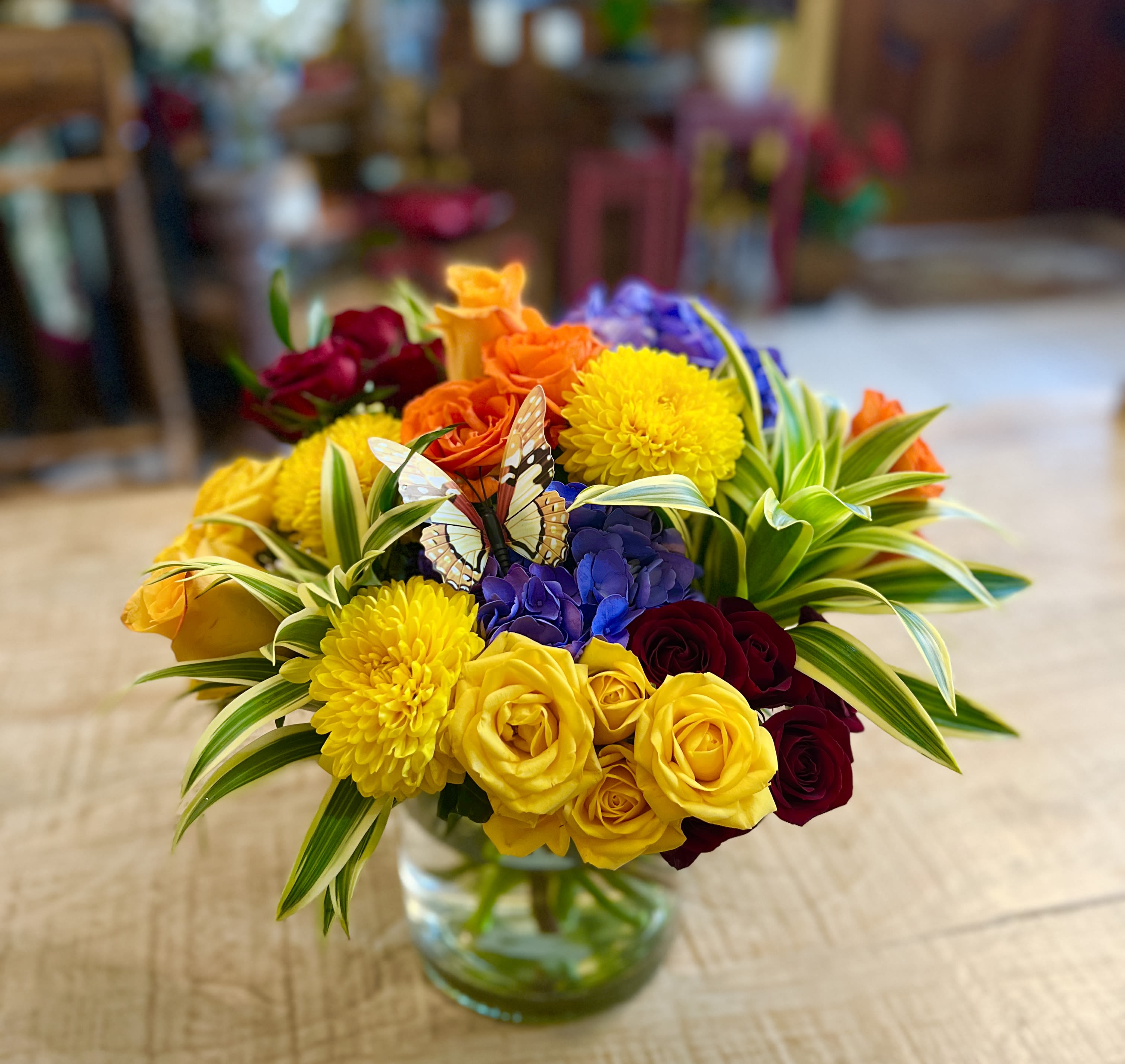 Summer Sensation - A bright, celebratory mix that makes someone feel truly seen-this Summer Sensation flower arrangement is a cheerful bouquet perfect for birthday bouquets, congratulations, or just because moments. The rounded, dense composition brings together yellow, red, orange, and purple blooms so the recipient is greeted with an immediate, joyful splash of color the moment they walk in the room.  Up front, golden yellow chrysanthemums and sunny roses create warmth and vibrancy, balanced by deeper red roses and blue‑purple hydrangeas that give the design contrast and visual depth. Variegated green-and-cream foliage frames the blooms for a fresh, garden‑picked look, and a decorative butterfly pick tucked near the center adds a whimsical, celebratory accent. Arranged in a clear glass vase, the compact shape works well on tabletops, desks, or as a centerpiece.  Ideal for sending to someone celebrating a milestone or to brighten an ordinary day, this bouquet reads as both festive and thoughtful-the kind of arrangement that will make the recipient smile and feel appreciated. Delivered ready to display in its vase; to keep it looking its best, refresh the water every couple of days and trim stems slightly when you change the water.