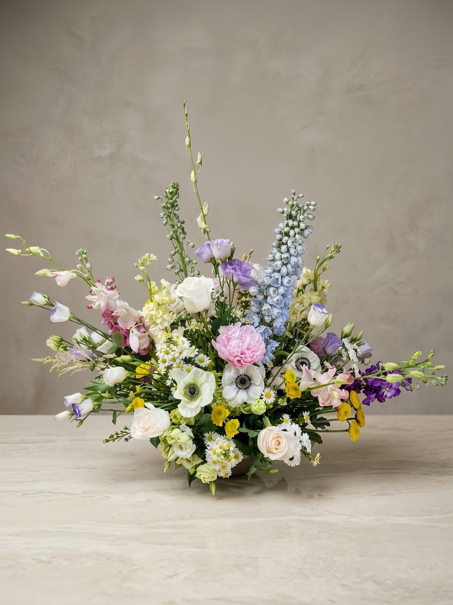 Bright Festive Centerpiece - A lush, elegant, and grand floral centerpiece beautifully crafted for first birthday banquets, weddings, and special parties. This vibrant arrangement features tall, majestic spikes of light blue delphinium and lavender stock, beautifully mixed with cream roses, bright yellow button poms, and elegant white anemones with dark centers. Designed to be full and multi-dimensional yet low enough for guest table conversations, it brings a cheerful and luxurious touch to any celebratory event.