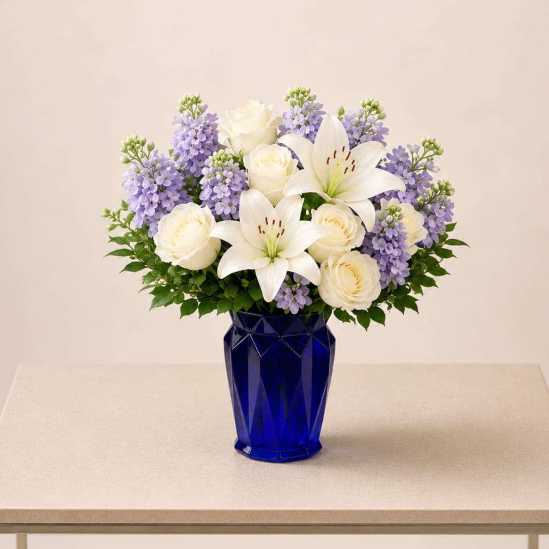 Serene Lavender Garden Arrangement - A refined and elegant floral arrangement featuring premium white roses, fragrant lavender stock, and graceful white lilies, beautifully designed in a modern cobalt blue vase. Accented with fresh seasonal greenery, this design offers a sophisticated balance of texture, color, and natural volume.  The combination of soft lavender tones and classic white blooms creates a timeless and calming aesthetic, making it an ideal choice for more meaningful and refined occasions.  Perfect for: sympathy, get well, thank you, thinking of you, and elegant everyday gestures.  Each arrangement is handcrafted using fresh, high-quality flowers. Bloom varieties may vary slightly based on seasonal availability while maintaining the overall style, color palette, and fullness.