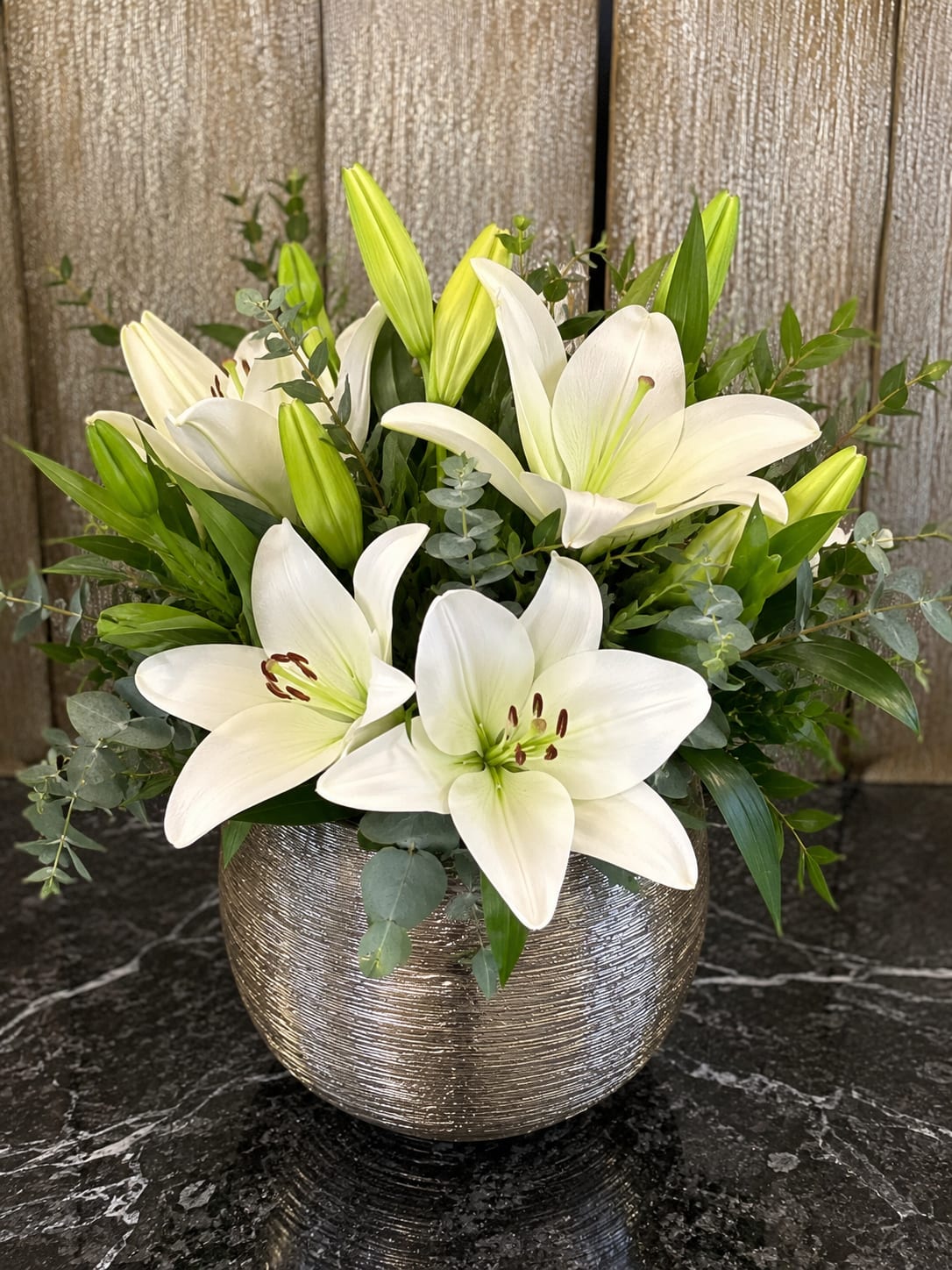 Moonlight Lilies - Pure white lilies open like moonlit blooms in this elegant arrangement-perfect as a sympathy tribute, an anniversary surprise, or a refined everyday gift. The lilies and their graceful unopened buds form a serene focal cluster that will make the person you're thinking of feel noticed and comforted, whether you mean sympathy, celebration, or quiet appreciation.  Crisp white petals are paired with airy, eucalyptus-like greenery for a fresh white-and-green palette that reads clean and contemporary. The stems are gathered in a rounded metallic vase with a textured silver finish, lending polished weight and modern contrast to the soft blooms. The composition is full and upright, with the lilies standing above the vase to create strong visual impact on a mantel, console, or bedside table.  This arrangement is designed to convey calm and elegance-a meaningful way to say you care. Standard care: keep in fresh water, trim stems every few days, and place out of direct sun to extend bloom life.