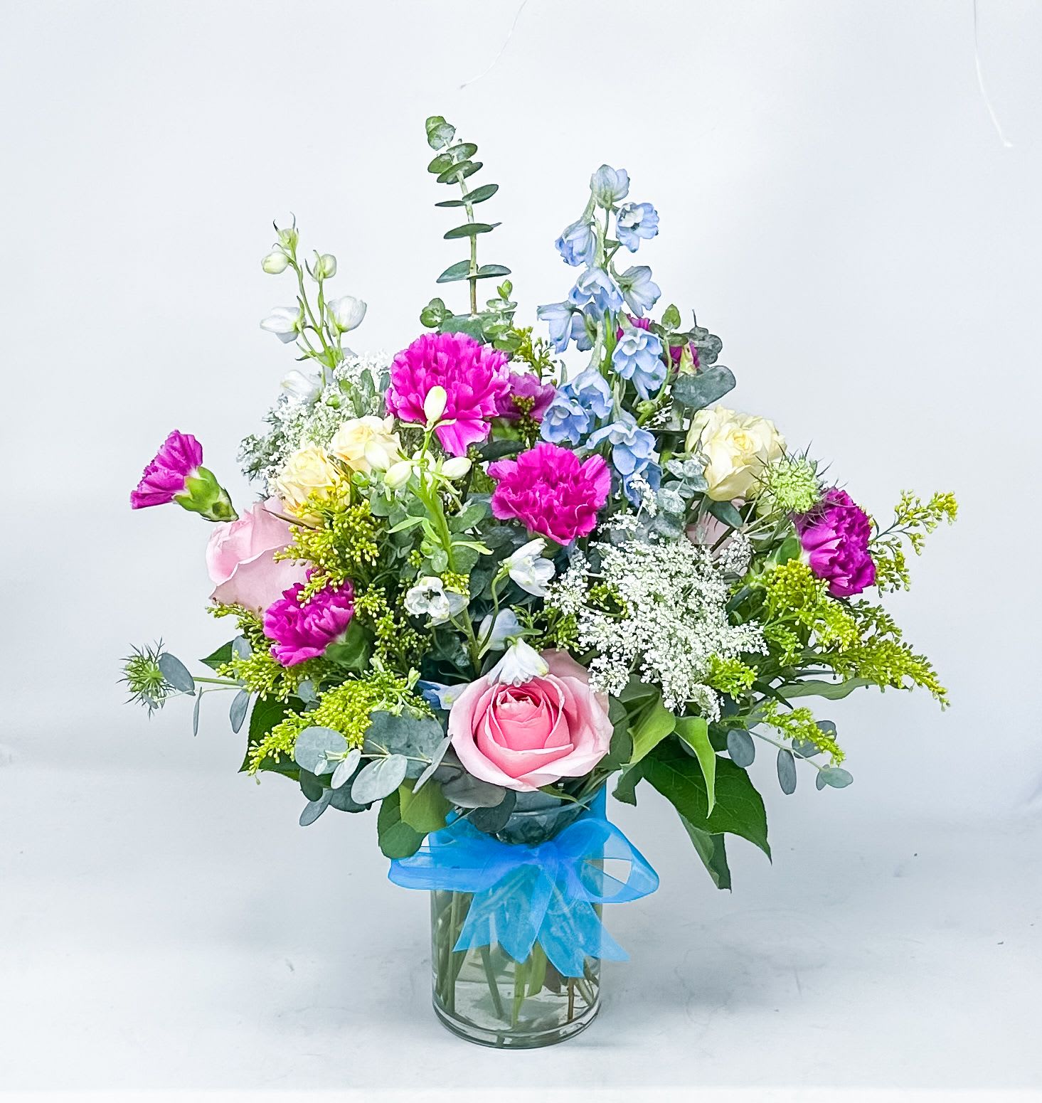 Mixed bouquet of pink, blue, and white flowers in a glass vase with a blue ribbon