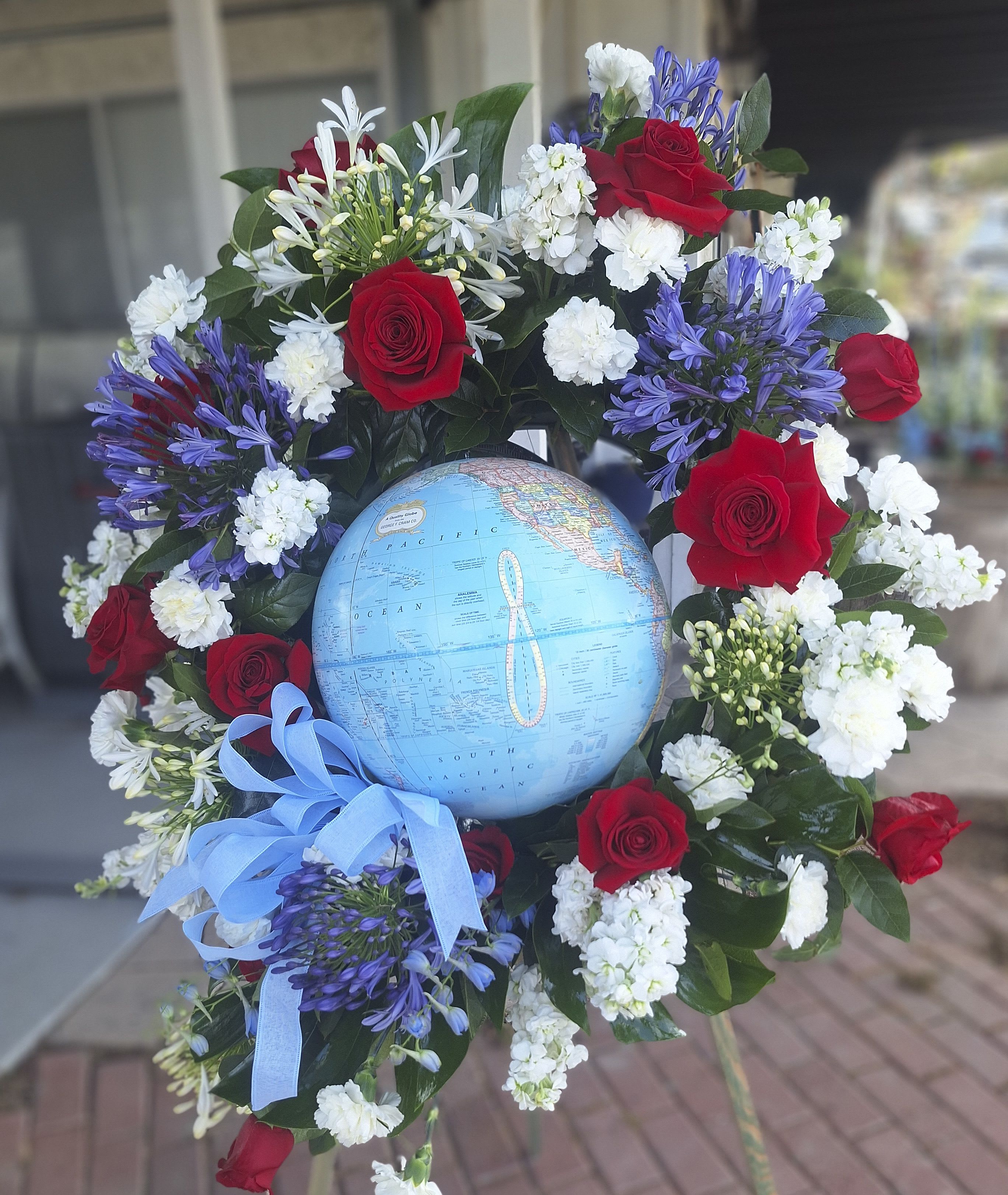 GLOBAL WREATH - A circular wreath of red, white, blue and purple blooms encircles a light blue globe, creating a dignified sympathy arrangement ideal for funeral or memorial services. The wreath format and globe centerpiece immediately read as a thoughtful tribute-perfect for honoring a loved one who had a connection to travel, service, or global work-while the red roses quietly convey respect and remembrance.  Clusters of red roses are evenly spaced around the ring, balanced by white carnations and small white filler flowers for soft texture, with purple blooms adding depth and contrast. A blue ribbon at the lower left echoes the globe's color and the piece arrives mounted on a standing easel for easy display. The overall size and scale work well as a focal memorial piece at a service.  Designed to make the recipient's family feel seen and honored, this arrangement offers a meaningful way to express sympathy and respect. Because it's assembled on an easel with secure hard goods, it's ready for immediate placement at a ceremony; standard care is to keep flowers cool and sheltered until the service.