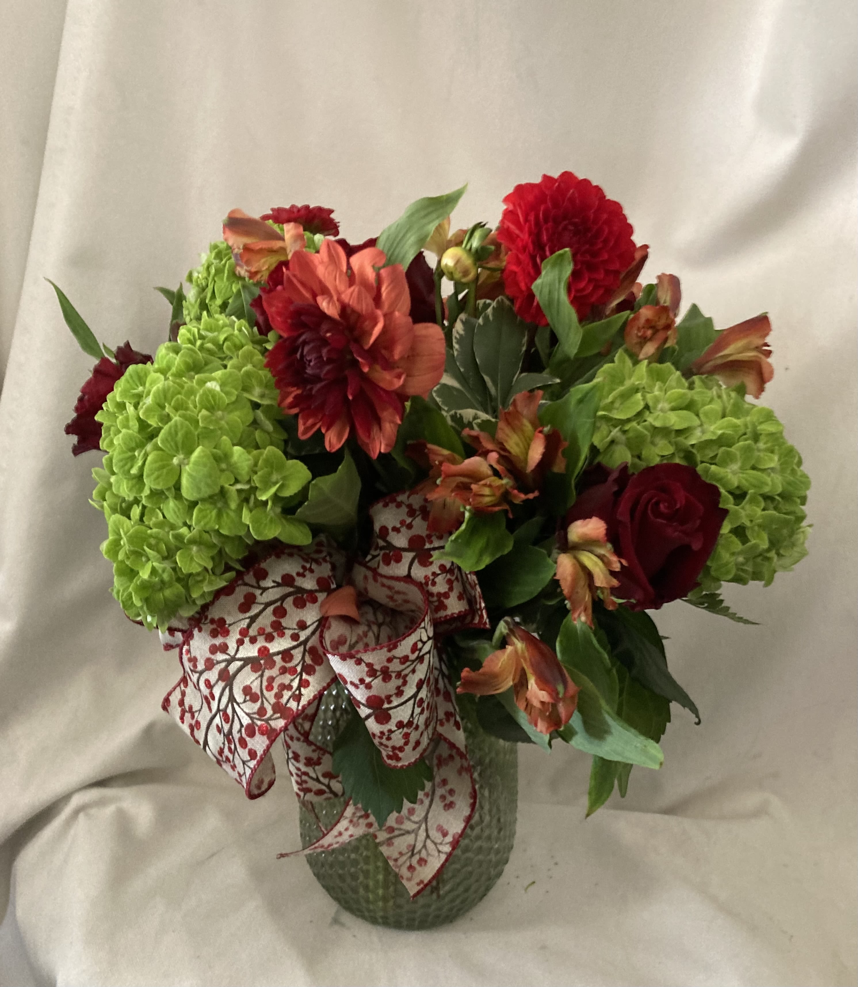 RED BEAUTY - Bright green hydrangeas anchor this bold RED BEAUTY flower arrangement, their lush, rounded clusters setting off deep red dahlias and a dark red rose for striking contrast-perfect as an anniversary roses gift or a festive birthday bouquet. Accents of orange alstroemeria and touches of white lift the palette so the person you're celebrating will feel both seen and delighted by the rich, layered color story.  This composition is arranged in a clear glass vase and finished with a white ribbon printed with red berries, giving the piece a subtly seasonal, decorative touch. Large hydrangea heads provide volume while the prominent red dahlia-like bloom and rose add focal drama; smaller orange alstroemeria weave texture and movement throughout. The result is a full, slightly formal presentation that works equally well as a table centerpiece or a thoughtful delivery to brighten someone's day.  Ideal for celebrations, special milestones, or as a decorative gift to bring warmth to a home, RED BEAUTY arrives ready to display. Keep flowers in fresh water, out of direct sunlight and away from heat sources to extend their life; contact us for same-day delivery options when you need a standout arrangement in a hurry.