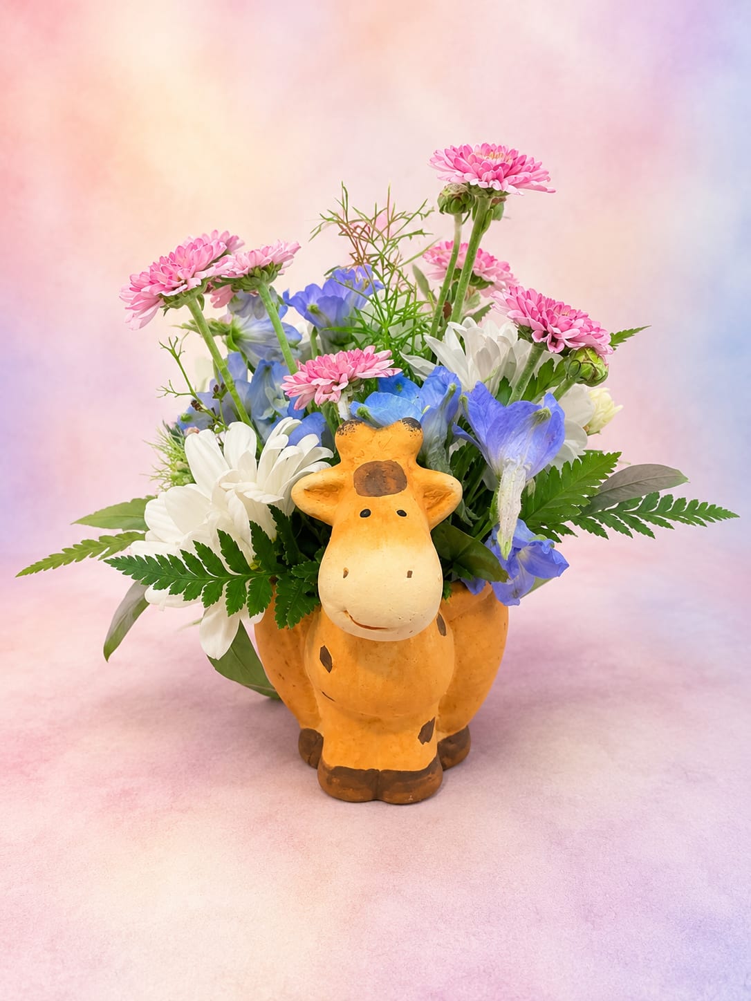 Safari Petals - Bring a smile with Pasture Petals, a charming mini arrangement designed in an adorable ceramic giraffe keepsake. Featuring cheerful pink mini daisies, crisp white daisies, vibrant blue delphinium, and wisps of airy greenery, this playful design captures the beauty of a blooming country meadow. Perfect for birthdays, get-well wishes, new babies, or just because, this sweet arrangement is a delightful gift that combines fresh blooms with a keepsake they'll enjoy long after the flowers have faded.