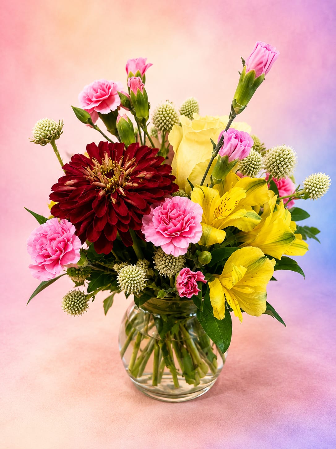 Berry Blossom Bliss - Description: Rich, vibrant, and full of garden charm, Berry Blossom Bliss is a joyful arrangement that pairs a striking burgundy zinnia with sunny yellow alstroemeria, soft pink carnations, buttery yellow roses, and whimsical globe thistle for a beautiful mix of color and texture. Designed in a clear glass vase with lush greenery, this fresh-from-the-garden bouquet has a naturally gathered look that's both elegant and cheerful. Perfect for birthdays, celebrations, thank-you gifts, or simply brightening someone's day, Berry Blossom Bliss is a delightful expression of happiness, warmth, and blooming beauty.
