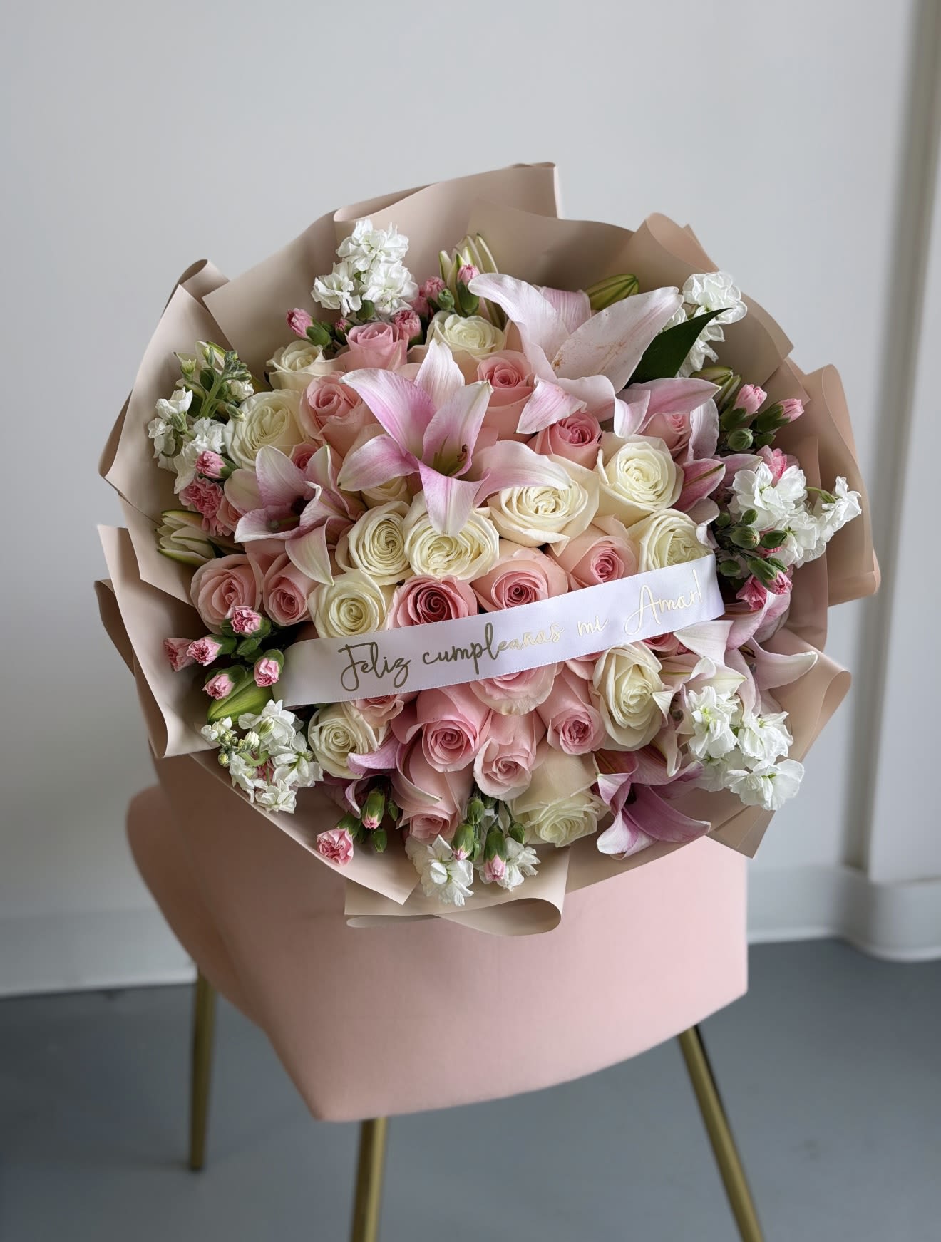 Blush Luxe 50 Rose Bouquet  - Soft pink and white roses gathered with pink lilies and delicate white-and-pink filler blooms create a lush, rounded hand-tied bouquet that reads as pure romance - perfect for a birthday bouquet, anniversary roses, or a "just because" surprise for the one you're thinking of. The pastel palette of pink, white, and soft peach feels tender and celebratory, making this arrangement a graceful choice for sending love, marking a milestone, or brightening someone's day for Mother's Day or a quinceañera.  The Luxe 50 Rose Bouquet features a generous mix of roses, lilies, and carnations arranged densely for full volume and soft texture. Wrapped in layered corean paper and tied with a white satin ribbon printed with a message, the presentation is both elegant and gift-ready; the paper gives a modern, tactile contrast to the petals while the satin ribbon adds a polished finish. The bouquet's rounded hand-tied shape is easy to hold and ideal for gifting in person or arranging in a vase at home.  Send this arrangement when you want her to feel truly celebrated and seen-whether it's a romantic anniversary, a heartfelt "thinking of you," or a milestone birthday. To keep the flowers fresh, place them in a clean vase with fresh water and trim stems at an angle every few days; our team prepares and secures each bouquet for same-day delivery when available so it arrives ready to impress.