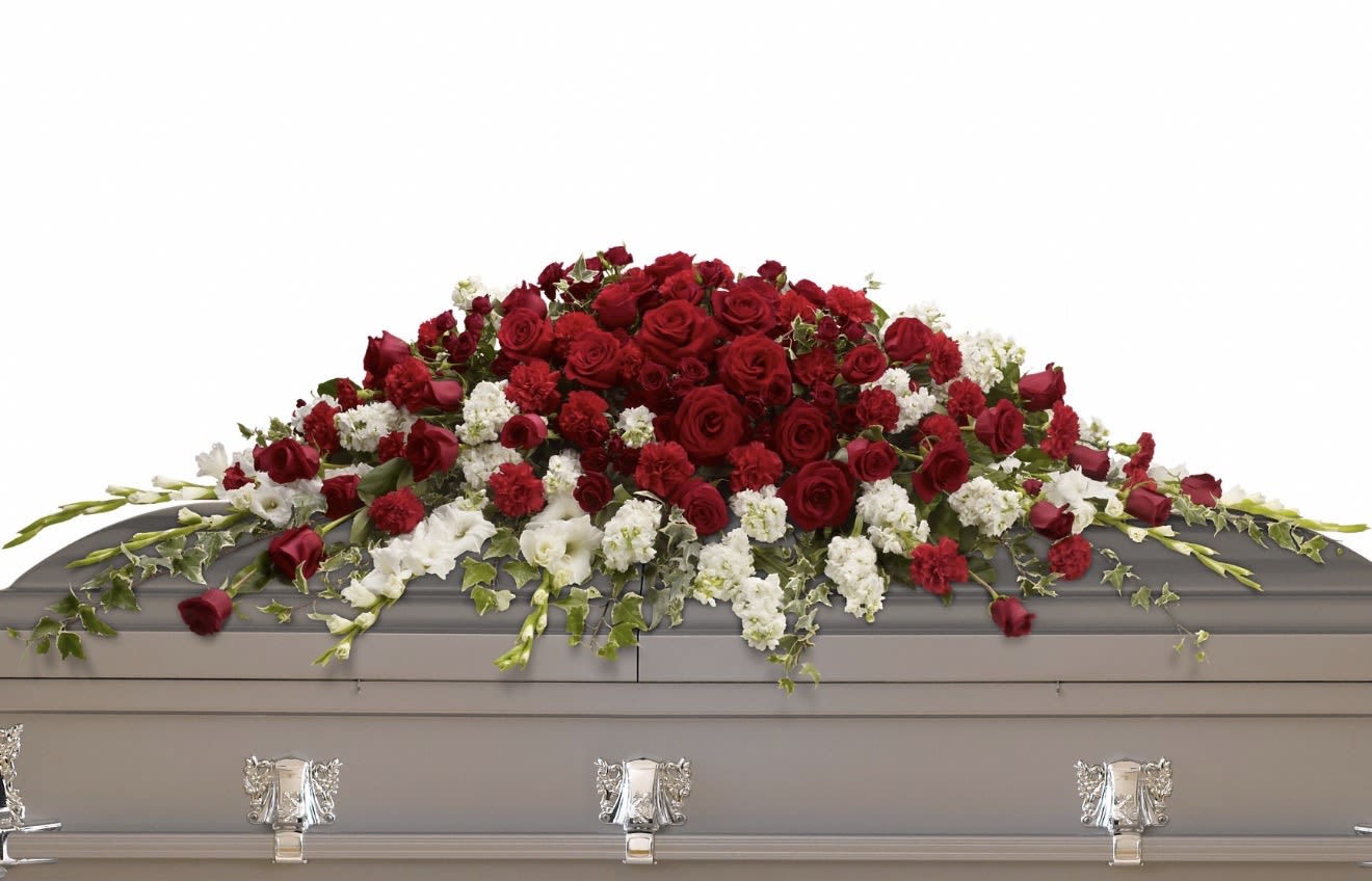 Garden of Grandeur Casket Spray - A dense mound of deep red roses and red carnations sweeps across the top of this funeral casket spray, offering a bold, respectful centerpiece for a memorial service. As a casket spray for sympathy and remembrance, the red-and-white palette conveys heartfelt love and solemnity-an appropriate tribute when words fall short.  White carnations are woven along the lower edge and contrasted with rich greenery and trailing ivy for a gentle cascade that softens the silhouette and adds quiet movement. Long-stemmed accent blooms extend outward to increase visual presence on the casket, creating a traditional, formal look that reads clearly from the front of the service.  Designed for funeral or memorial use, this large spray is sized to cover the top of the casket and present a unified, elegant display at visitation or graveside. Minimal care is needed from the recipient-arrangement is delivered ready to place-so the flowers arrive fresh and prepared to honor the person you're remembering.