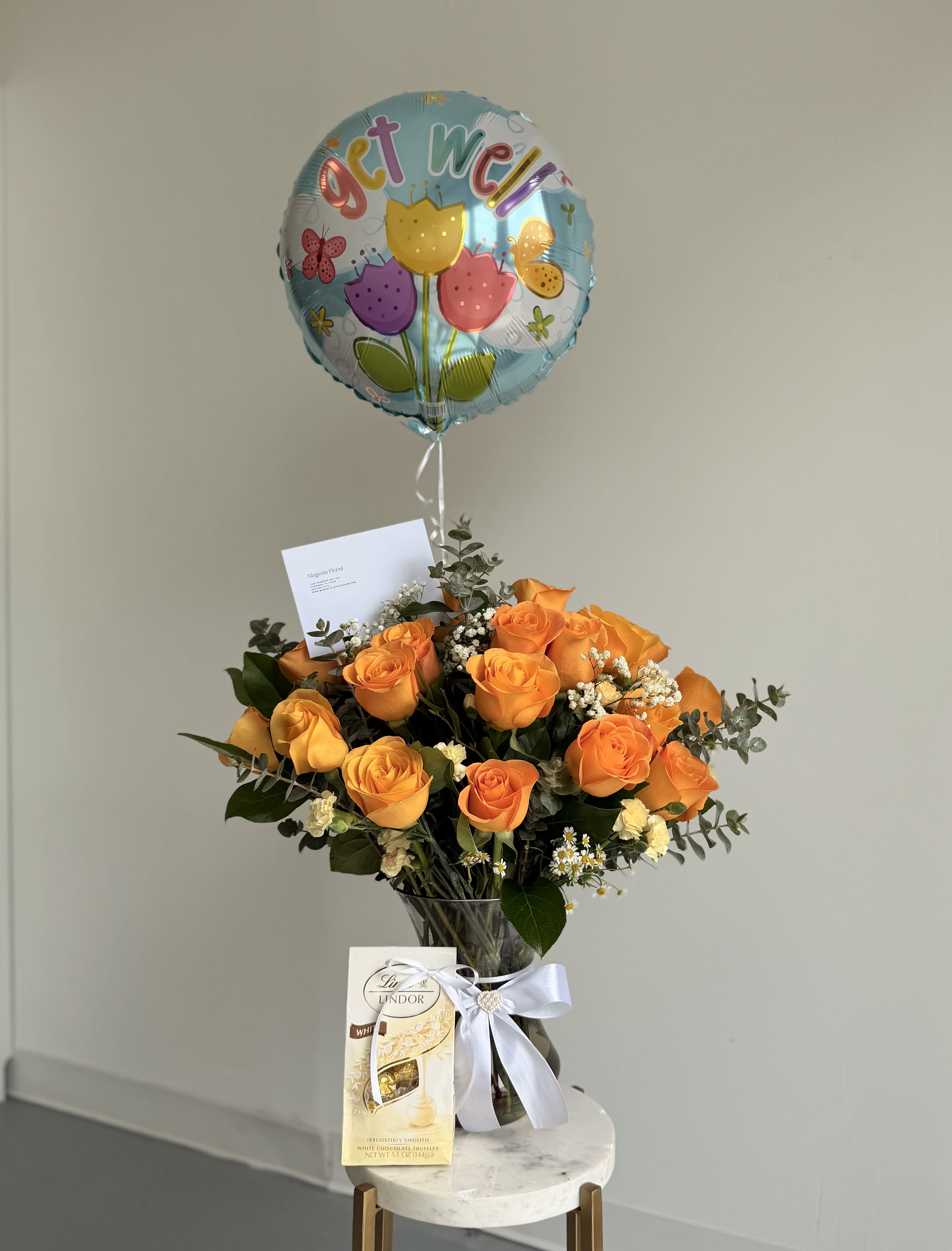 Golden Bloom Gift Set - A bright yellow roses takes center stage in this flower arrangement, sending a warm, celebratory message for get well, birthdays, congratulations, or a thoughtful "just because" surprise. The Golden Bloom Gift Set feels cheerful and uplifting-perfect for saying thank you, cheering someone on during recovery, or adding a romantic, sunny touch to an anniversary or sweet note of love.  The bouquet sits in a clear glass vase tied with a crisp white ribbon, with yellow roses softened by small white filler blooms and airy green foliage that adds lightness and texture. A  foil balloon floats above for instant festivity, and a bag of Lindor chocolates is added to create a complete gift set that delights both the eyes and the palate.  Ideal for the person who deserves to feel seen and celebrated, this arrangement works as a get-well pick-me-up, a romantic surprise, or a generous thank-you. Vase-ready and thoughtfully finished, it arrives ready to display-no fuss, just a bright, fragrant moment that lets them know you care.