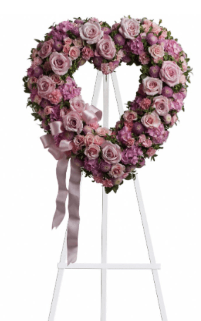 Rose Garden Heart - A heart-shaped standing wreath crafted in soft pinks, lavenders and clean whites-designed to offer a tender, timeless tribute for sympathy and funeral services. The Rose Garden Heart arrangement places roses front and center for a romantic, comforting presence, while the heart silhouette speaks plainly to love and remembrance for the person you're honoring.  Densely packed roses are paired with spray roses, carnations and chrysanthemums to create layers of texture and gentle color variation; small clustered blossoms and round purple accents add depth across the wreath. A satin ribbon drapes from the lower left for a ceremonial finish, and the piece is displayed on a white easel for an elegant, upright presentation.  Perfect for family members, dear friends, or as a memorial from a community, this heart wreath communicates lasting affection and sympathy at the service or graveside. The arrangement arrives ready to place on the included easel; to keep it looking its best, display in a cool, shaded area away from direct sun and wind until the service.
