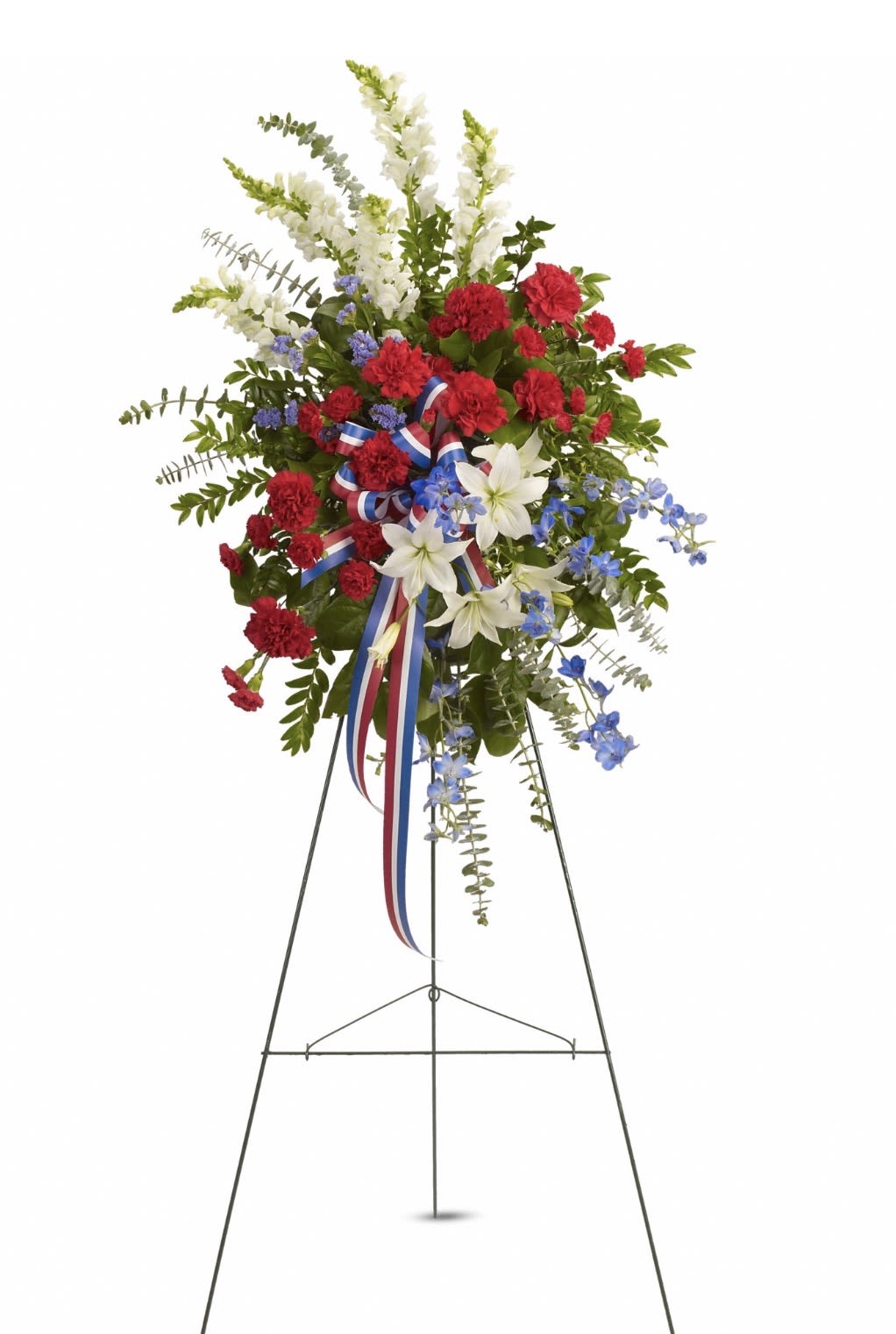 Sacred Duty Spray - Clusters of deep red carnations and serene white lilies give this standing spray a dignified, ceremonial presence-perfect as a sympathy arrangement or for a funeral tribute. The tall, asymmetrical design carries airy white spikes and blue delphinium accents that lift the composition, while a red, white, and blue ribbon adds a respectful, patriotic touch for memorials that honor service or national remembrance.  Designed to stand on a metal easel, the spray is full and layered with long stems and textural snapdragons woven through the main body to add movement and depth. Colors read clearly from a distance-rich red, crisp white, cool blue and verdant greenery-so the person you're honoring will be remembered with a composed, thoughtful display. Care: assembled fresh for delivery and intended for standing display; keep out of direct heat and mist sparingly if needed to preserve bloom life.