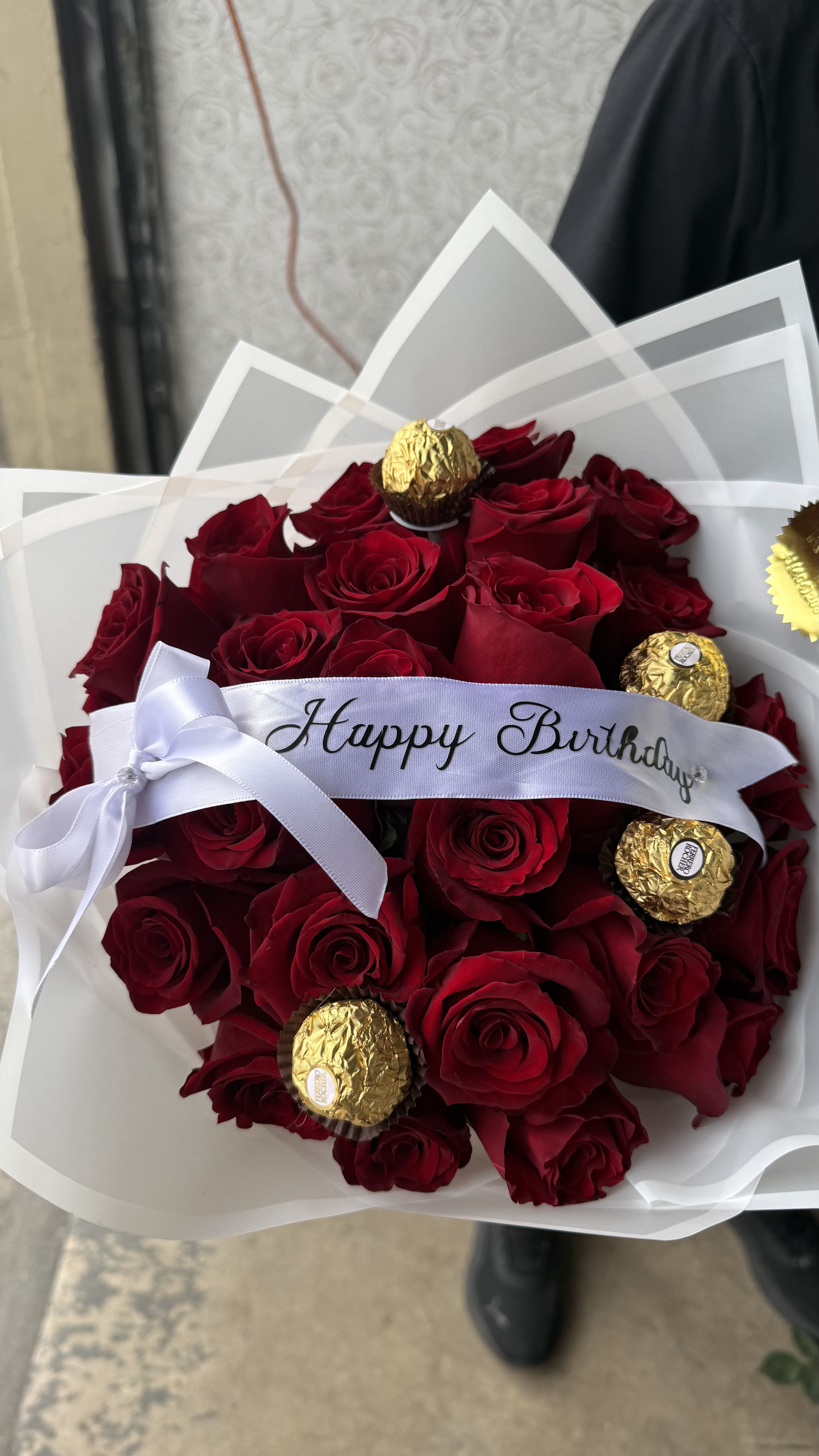 Red Roses - Ferrero Bouquet - Tightly clustered deep red roses form a lush, rounded birthday bouquet that reads as both romantic and celebratory - a perfect anniversary or birthday gift for the person you want to make feel truly cherished. The arrangement's compact shape and rich crimson petals create an immediate visual impact that says "I thought of you" the moment it arrives.  Interspersed among the red roses are several gold-wrapped Ferrero Rocher chocolates, adding a touch of indulgent surprise and contrasting texture. A white satin ribbon across the front with "Happy Birthday" and a clean white paper wrap finish the look for a gift-ready presentation. Colors here - deep red roses with bright gold accents and white trim - keep the feel classic and elegant.  Ideal for birthdays, anniversaries, or any romantic celebration, this bouquet sends a clear message of affection and celebration to the recipient. The compact, hand-tied style travels well; to keep the roses looking fresh, place them in water on arrival and recut stems at an angle if you transfer them to a vase.