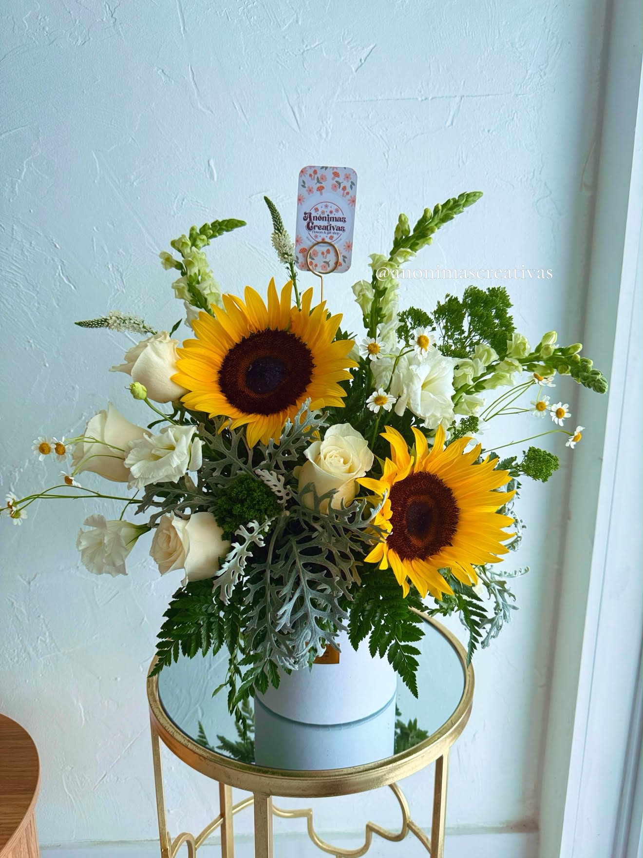 Sunflower Serenity Arrangement - Celebrate life’s brightest moments with this fresh and elegant floral arrangement featuring radiant sunflowers, classic white roses, lisianthus, snapdragons, chamomile, and lush seasonal greenery. Designed in a modern white keepsake container, this cheerful arrangement blends warmth, elegance, and natural beauty.  Perfect for birthdays, thank-you gifts, congratulations, housewarmings, or simply to brighten someone’s day. Handcrafted by our expert florists using premium fresh flowers, it’s a thoughtful gift that leaves a lasting