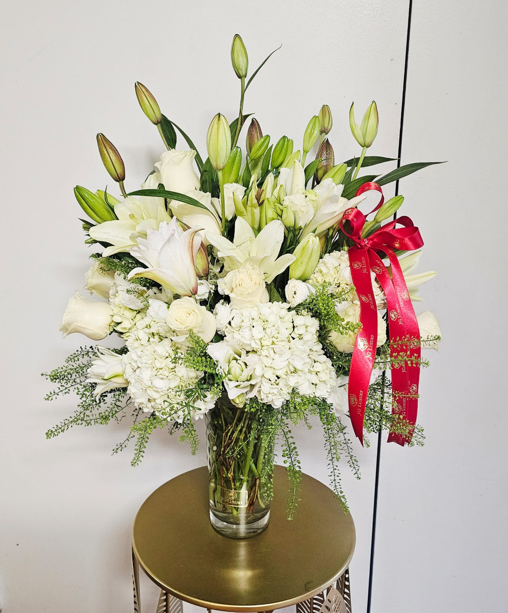 Lily Love - featuring elegant white lilies, premium white roses, lush hydrangeas, and delicate seasonal blooms, accented with fresh cascading greenery. Designed in a clear glass vase and finished with a luxurious satin ribbon, it brings a sense of peace, sophistication, and effortless beauty to any occasion. Perfect for celebrating life's special moments or expressing heartfelt sympathy with grace and elegance.