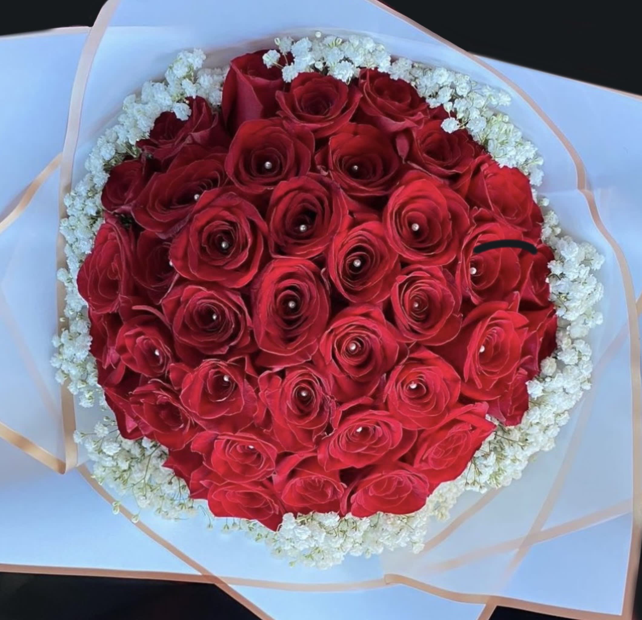 Round bouquet of red roses with pearl centers, edged with small white flowers in white wrapping.