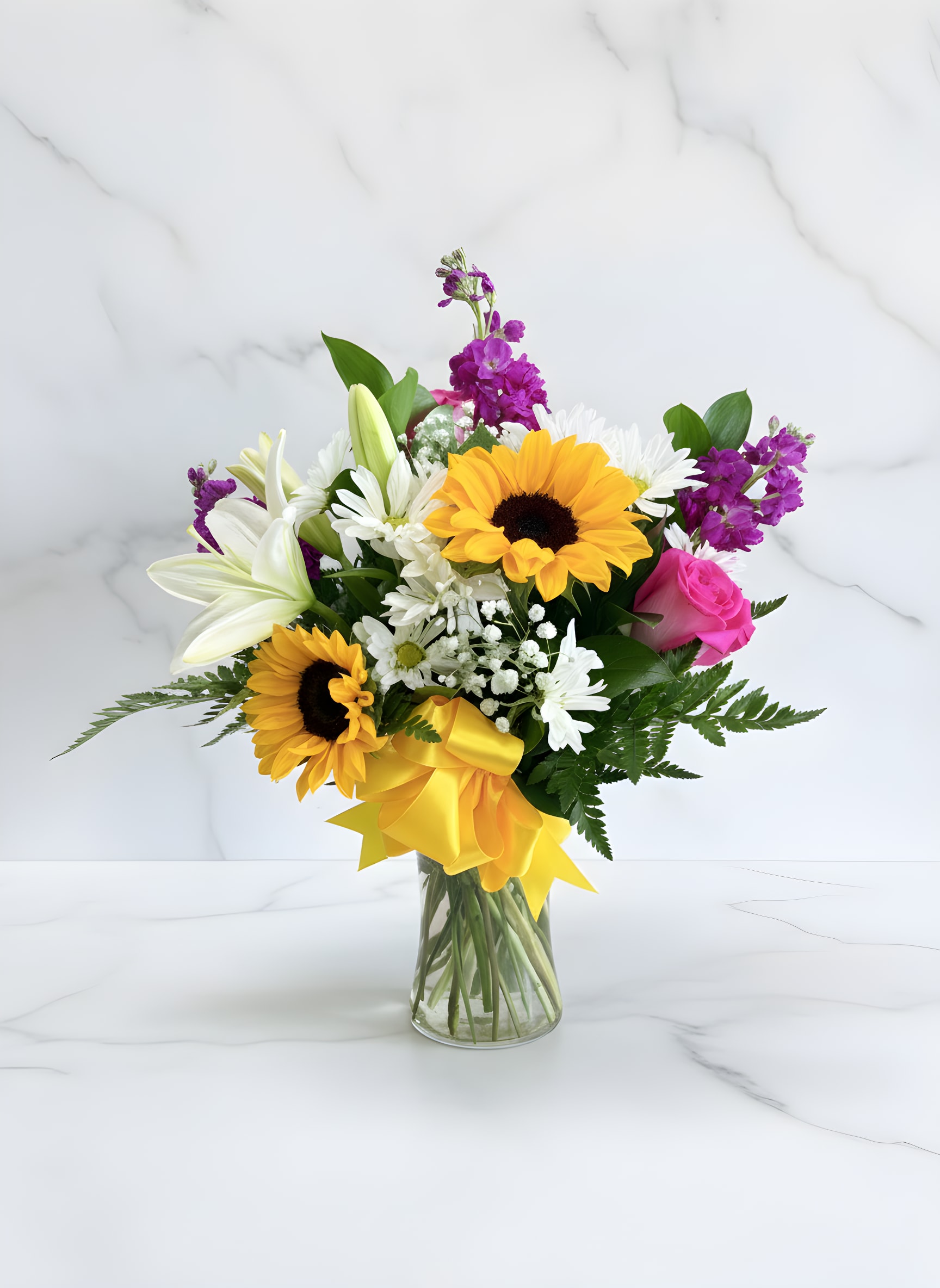 Bella Girasole - Make every celebration unforgettable with Bella Girasole, an elegant floral arrangement designed in a classic clear glass vase and finished with a beautiful yellow satin ribbon. Featuring radiant sunflowers, fragrant white lilies, premium pink roses, fresh white daisies, purple stock, baby's breath, and lush greenery, this stunning bouquet brings joy and sophistication to any occasion. Perfect for birthdays, anniversaries, Mother's Day, congratulations, thank-you gifts, get well wishes, new baby celebrations, or just because. Handcrafted with premium fresh flowers and designed to leave a lasting impression.