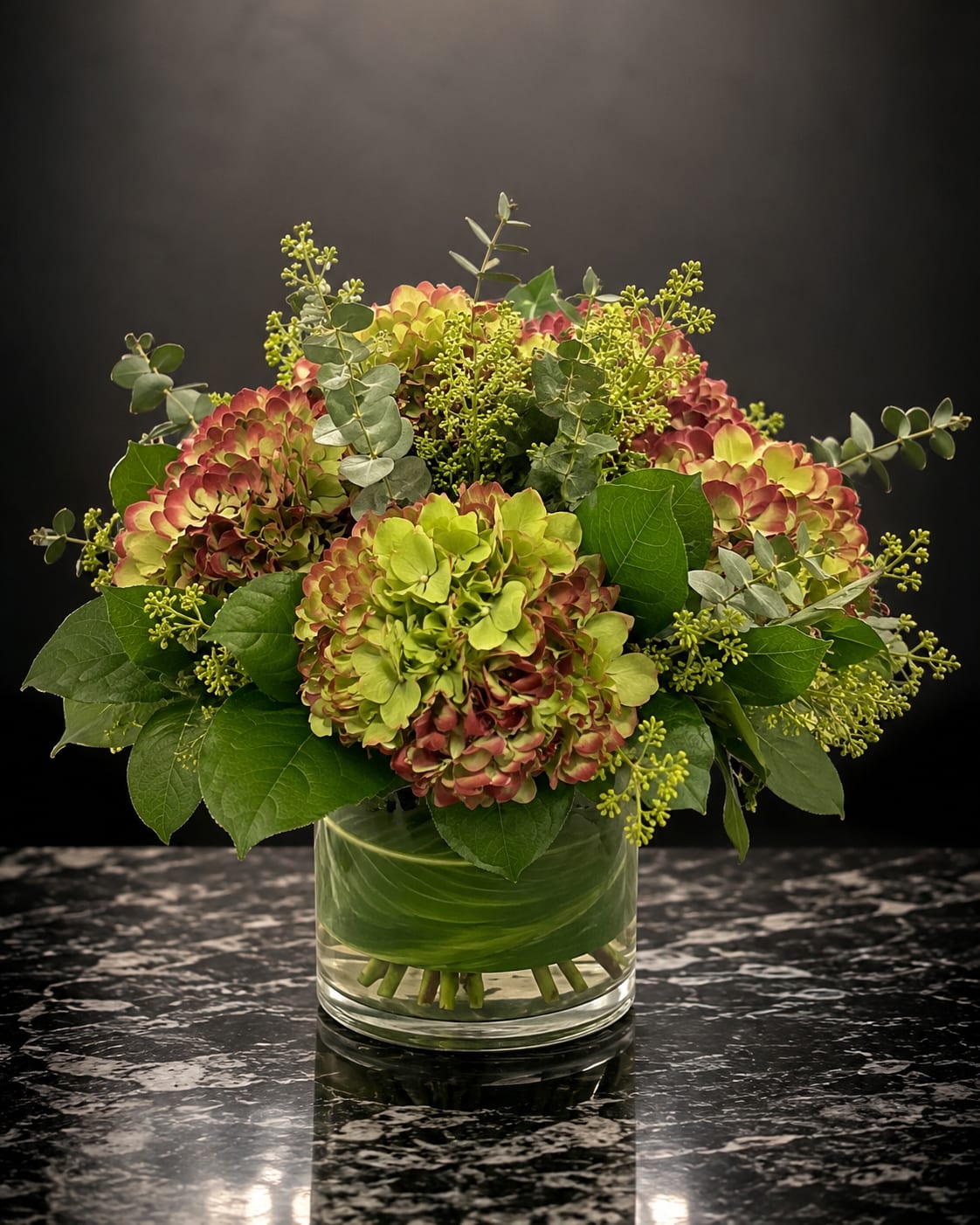 Velvet Hydrangea - Lush clusters of green, pink, and muted purple hydrangeas form a full, rounded bouquet that reads as both elegant and effortlessly garden-fresh - perfect as a birthday bouquet, anniversary flowers, or a thoughtful "just because" gift. The soft, velvety texture of the hydrangea heads and the gentle color transitions create an immediate sense of warmth; the person you send this to will feel seen and celebrated by its generous, romantic presence.  This arrangement features several large hydrangea blooms gathered with broad leafy foliage and airy branching greenery for contrast and texture. Stems are arranged in a clear cylindrical glass vase so the natural stems show through, giving a clean, modern look that suits tabletops, entryways, or office desks. Sized to make a notable impression without overwhelming a space, it offers long-lasting freshness typical of hydrangeas - change the water regularly and trim stems for best enjoyment.