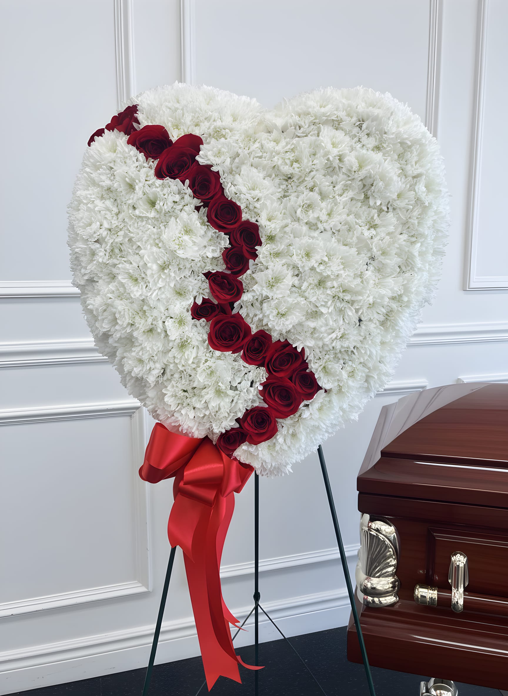 Eternal Love Broken Heart Standing Spray - Honor a cherished life and express heartfelt sympathy with our Eternal Love Broken Heart Standing Spray. This elegant funeral tribute features a heart-shaped design crafted with lush white chrysanthemums, symbolizing peace, purity, and everlasting remembrance. A striking diagonal ribbon of premium red roses gracefully divides the heart, representing enduring love, deep devotion, and a bond that lives on forever.  Displayed on a standing easel with a luxurious red satin bow, this sympathy arrangement offers a timeless expression of love and remembrance. It is a beautiful choice for funeral services, memorial ceremonies, celebration of life services, or graveside tributes.