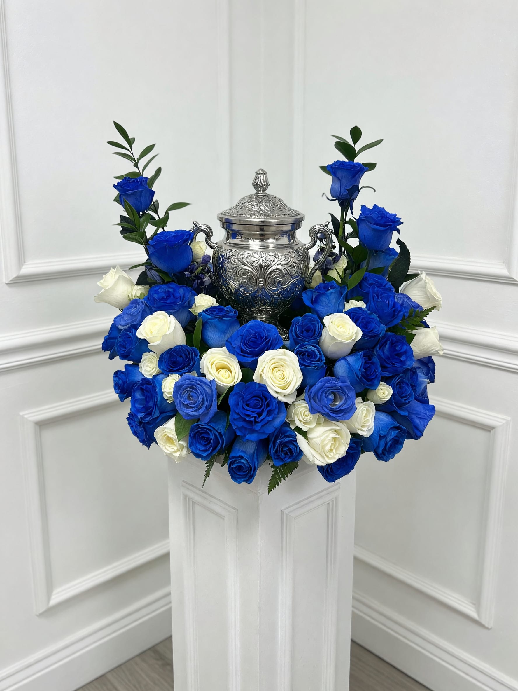 Eternal Honor Cremation Tribute - Celebrate the life and legacy of a loved one with our Eternal Honor Cremation Tribute. This elegant urn arrangement is handcrafted with stunning royal blue and pure white roses, beautifully surrounding the cremation urn to create a meaningful and dignified display. The striking contrast of vibrant blue roses and pristine white roses symbolizes loyalty, peace, remembrance, and everlasting love.  Designed to serve as the focal point of a funeral, memorial service, or celebration of life ceremony, this luxurious floral tribute offers a heartfelt expression of sympathy while honoring treasured memories. Lush greenery adds natural beauty and depth, creating a timeless arrangement that reflects grace, respect, and devotion.