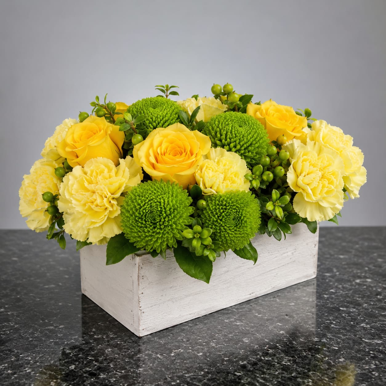 Emerald Sunshine - Bright yellow roses and ruffled yellow carnations combine with vivid green chrysanthemum pompons to create a cheerful spring bouquet perfect as a birthday bouquet, thank-you arrangement, or just-because gift. The bright, sunny palette of yellow, green, and touches of white reads joyful and polished - the kind of floral gift that makes the person you're thinking of feel instantly uplifted.  Flowers are gathered low in a clean, modern white wooden box that keeps the design compact and easy to display on a desk, countertop, or dining table. Small green berry accents weave through the roses, carnations, and mums to add texture and a fresh contrast to the warm yellow blooms. The low rectangular format highlights the rounded shapes of the flowers and makes the arrangement feel abundant without towering.  Ideal for spring celebrations, housewarmings, or as a bright thank-you gesture, this arrangement is designed for immediate impact and easy styling in any room. The sturdy white wooden box arrives ready to display - no vase required - and the mix of long-lasting carnations and mums with classic roses helps ensure the recipient enjoys the sunny look for days.