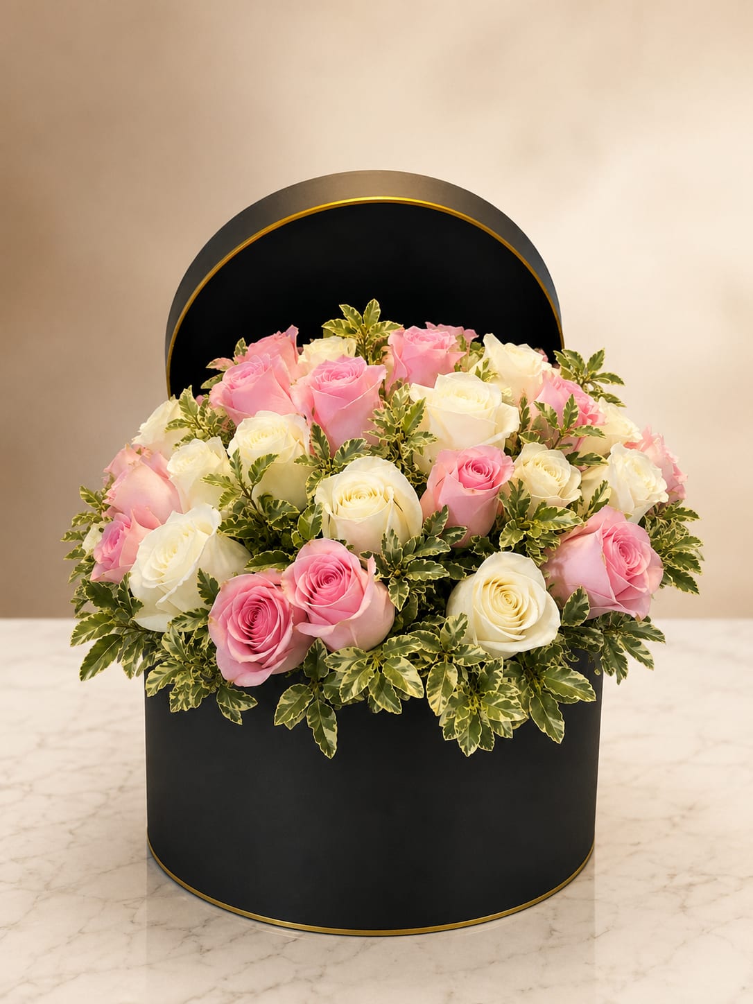 Soft Embrace - Soft pastel roses arranged in a chic black round hat box with gold trim create a polished, romantic look that reads both modern and timeless - perfect as a birthday bouquet, anniversary roses, or an everyday expression of love and appreciation. The contrast of pink and white blooms against the dark container makes the recipient feel cherished and seen, the kind of arrangement that quiets a busy day and makes someone smile.  A dense cluster of pink and white roses fills the hat box, surrounded and softened by variegated green foliage that spills gently over the rim for a full, rounded silhouette. The black box with subtle gold edging adds elegant contrast and keeps the design feeling contemporary while the lush texture of the roses remains the focal point.  Send this to mark a romantic milestone, celebrate a loved one, or say "I'm thinking of you" with effortless refinement. Standard sizing creates a generous tabletop statement piece; keep roses fresh by changing water and trimming stems if you transfer them to a vase, or enjoy the boxed presentation as an immediate, low-maintenance gift.