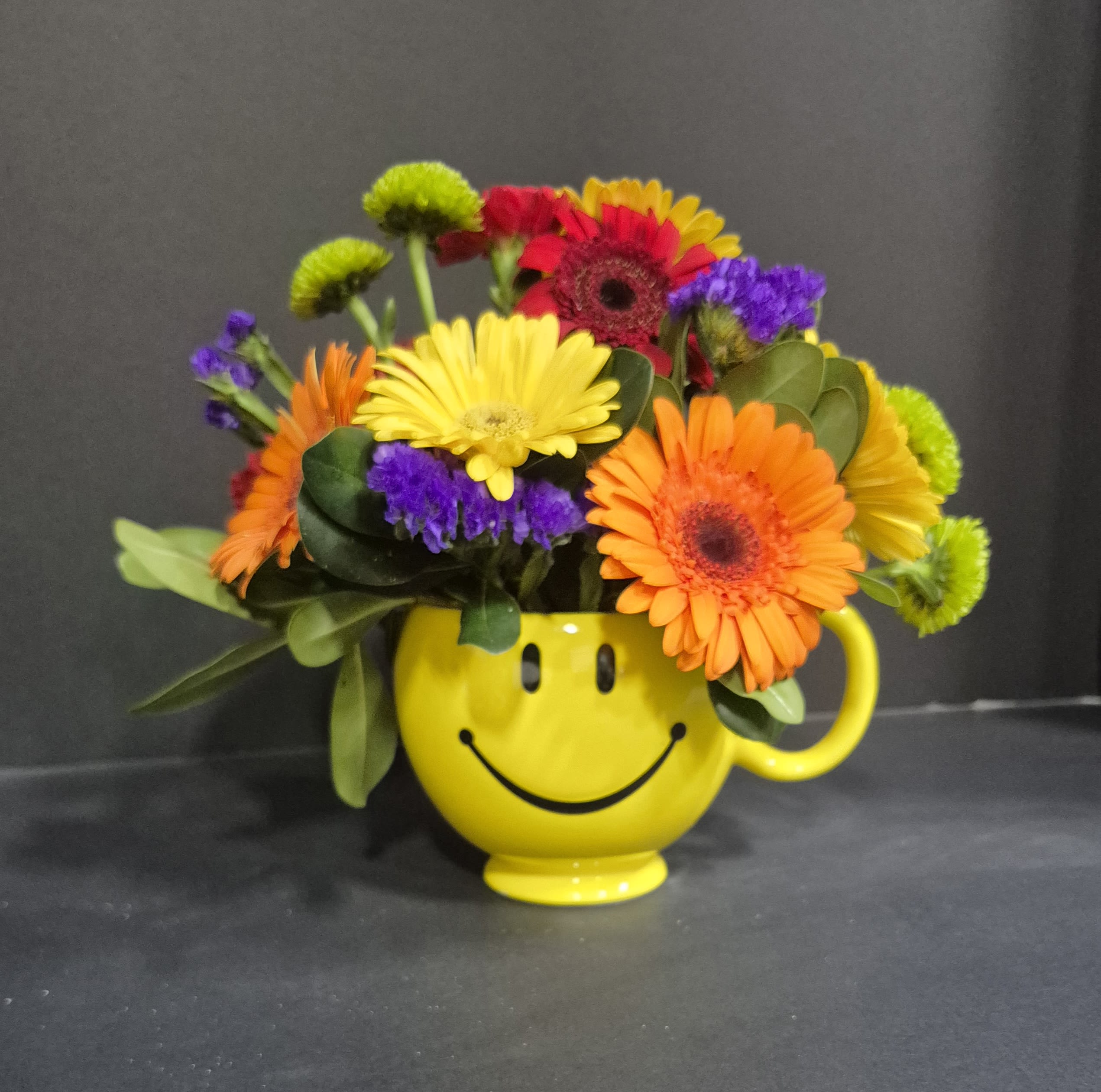 Sending Bright Smiles - A bright, colorful seasonal mix of flowers in a smiley face mug.  Send your loved one a burst of happiness for any occasion or just because. Call directly if you have any specific color requests.