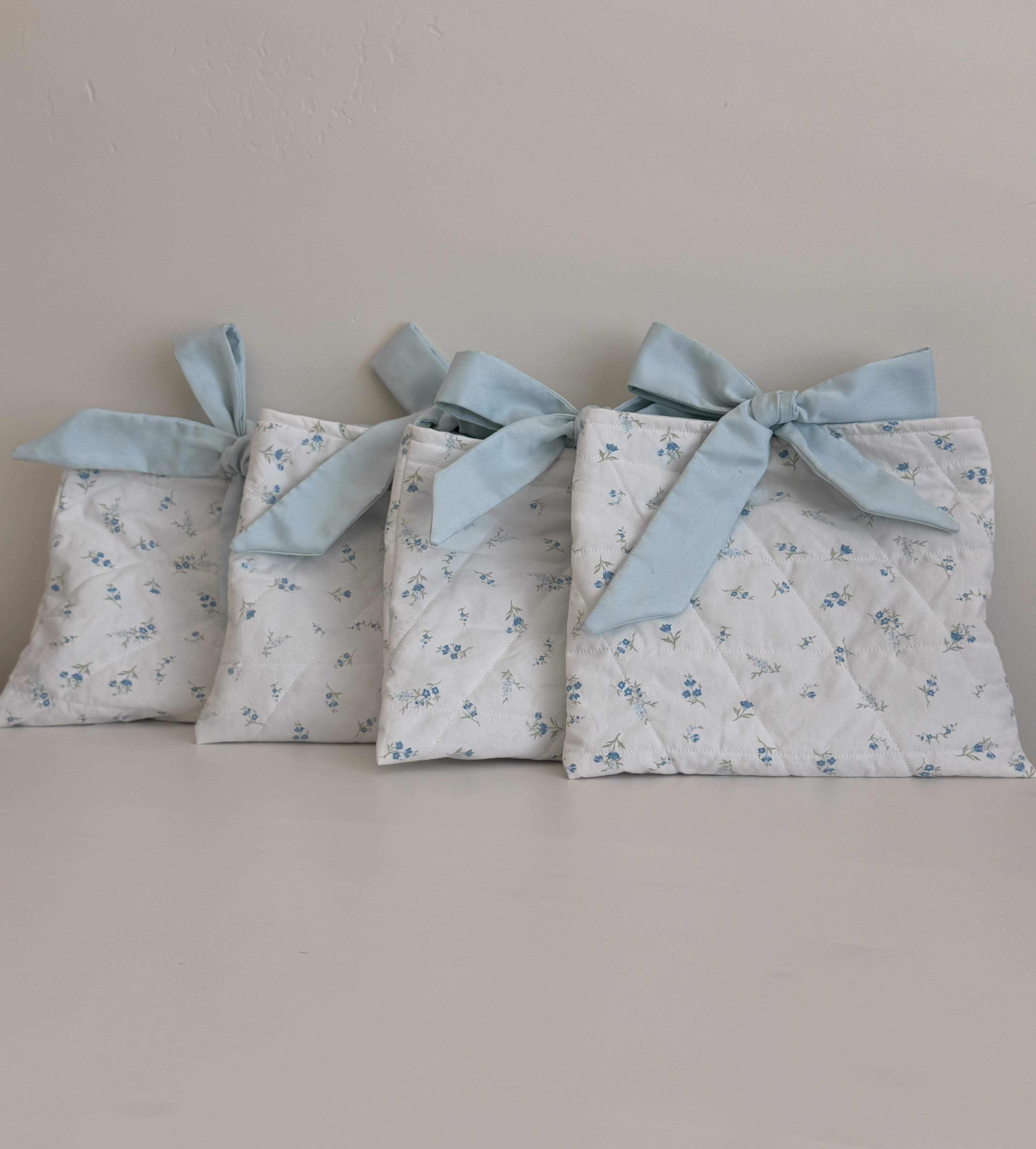 Hand Quilted bag - Delicate hand-quilted fabric pouche make a charming alternative gift presentation for a baby shower, bridal party, or thoughtful hostess gift. This white-and-blue set reads like a vintage-inspired keepsake-perfect for a small, intimate occasion where presentation matters. The soft palette and floral print create a calm, sweet impression that will make the person you're thinking of feel noticed and cherished.  Each of the four pouches is made from quilted white fabric patterned with a tiny blue floral print and finished with a pale blue ribbon tie at the top. Sized to hold small treats, notes, or a petite favor, these soft pouches arrive arranged in a neat row and can be used as decorative accents, party favors, or reusable gift packaging. Gentle spot cleaning will preserve the fabric and ribbon; if you need help selecting small florals or gifts to tuck inside, we can recommend pairing options to suit baby showers, bridesmaids' gifts, or host/hostess presents.  Can be used as a small diapr bag or as a bag for your Kindle.