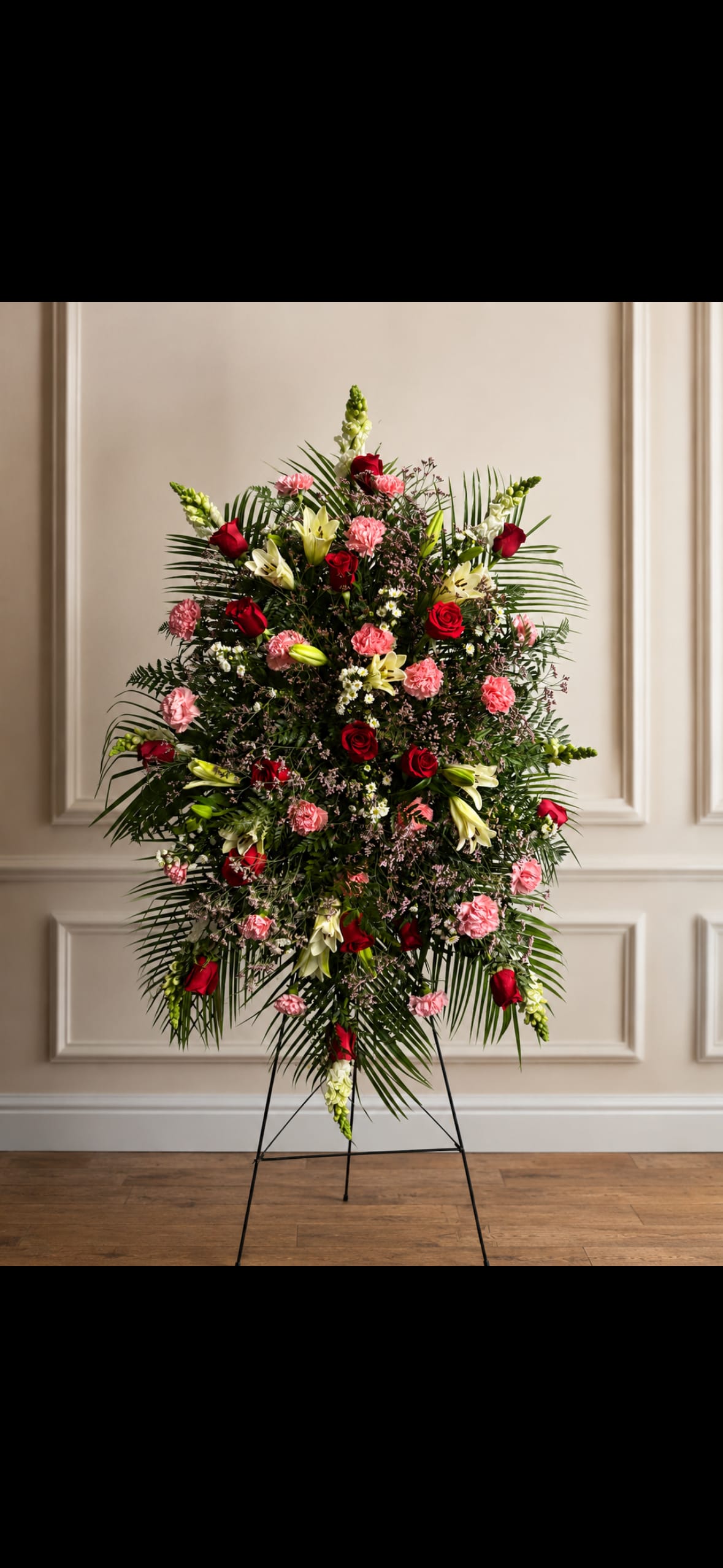 Red White and Pink Easel - A large standing spray of red, pink and white blooms designed to offer a graceful, ceremonial statement for sympathy or memorial occasions. The arrangement's strong vertical height on a sturdy metal easel creates a formal silhouette that honors the person you're remembering, while the saturated red roses paired with soft pink carnations and pale lilies bring warmth and quiet beauty to the space.  This piece layers roses, carnations, lilies, snapdragons and airy filler flowers into a dense, rounded composition accented with long palm fronds for texture and presence. The overall palette-deep red, blushing pink, crisp white and lush green-reads classic and elegant from every angle; it arrives ready to display on the included metal easel. Sized for visibility at services and viewings, it's arranged for lasting vase life with standard floral care: keep cool, away from direct sun, and hydrate any water source provided per your order instructions.