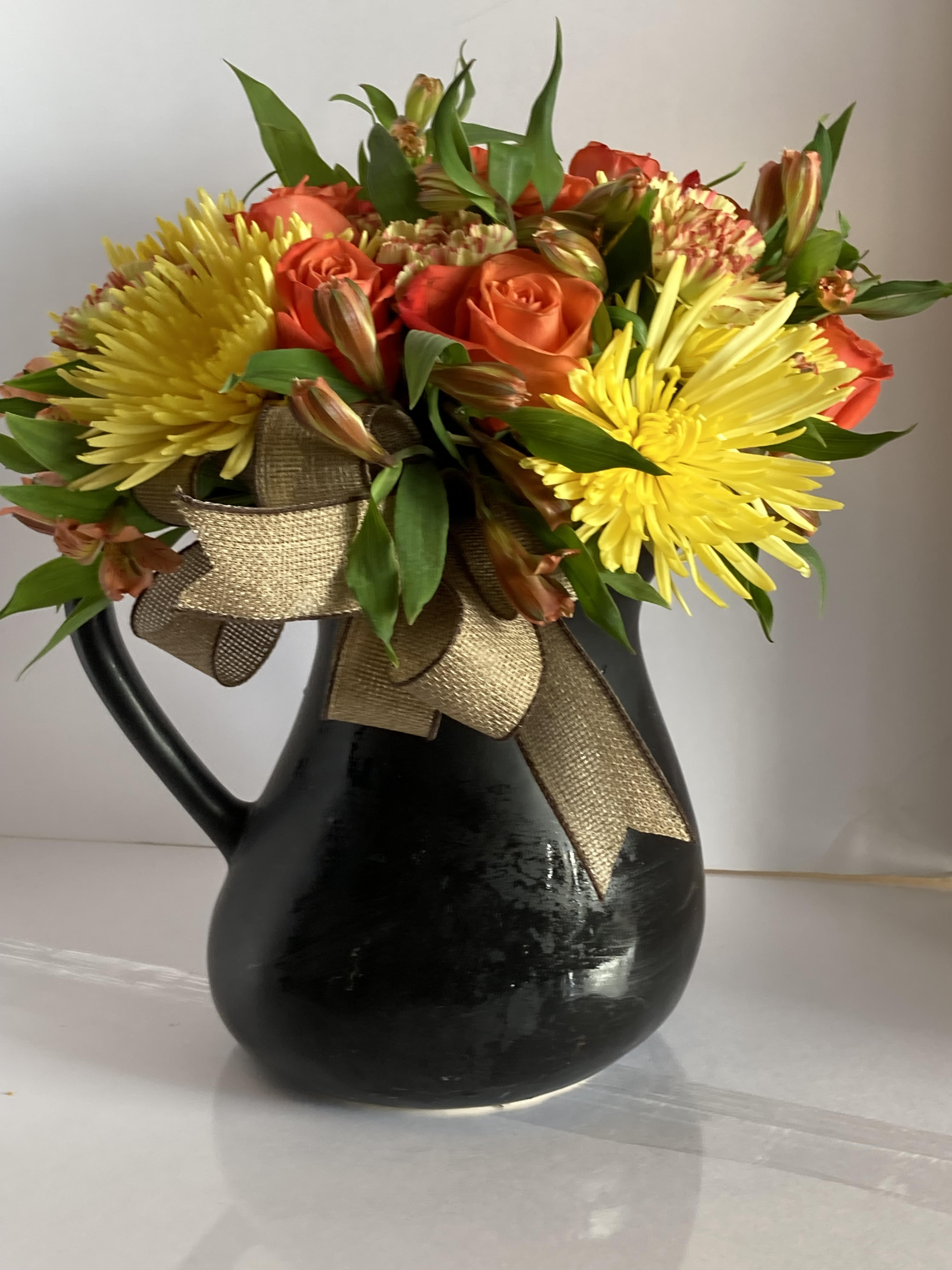 Orange roses and yellow chrysanthemums in a black pitcher vase with a gold ribbon
